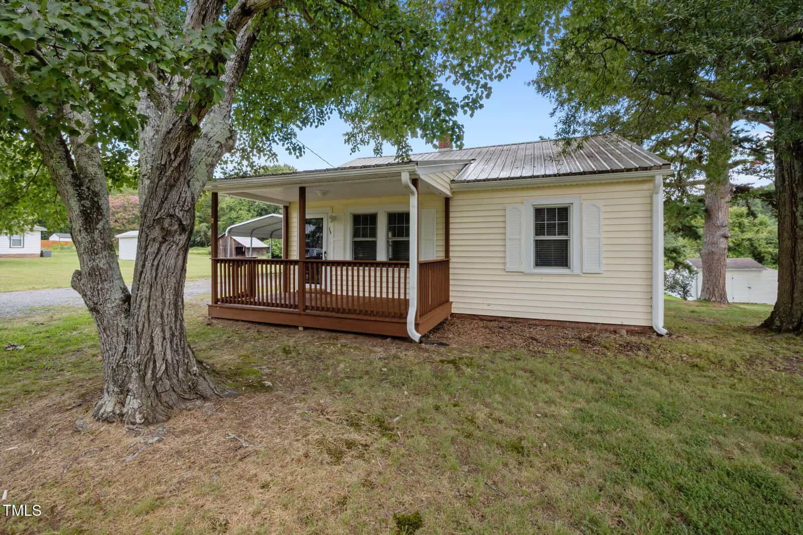 A small, single-story yellow house with white trim, a covered wooden front porch, and a metal roof is surrounded by grass and shaded by leafy trees. A gravel driveway is visible to the left.