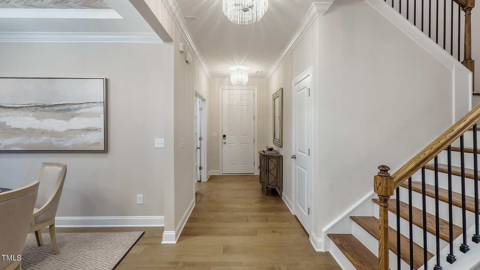 A bright hallway with light-colored walls and wood flooring, featuring a staircase with wooden steps and black railings, a chandelier, artwork, a small table with a mirror, and a white front door.