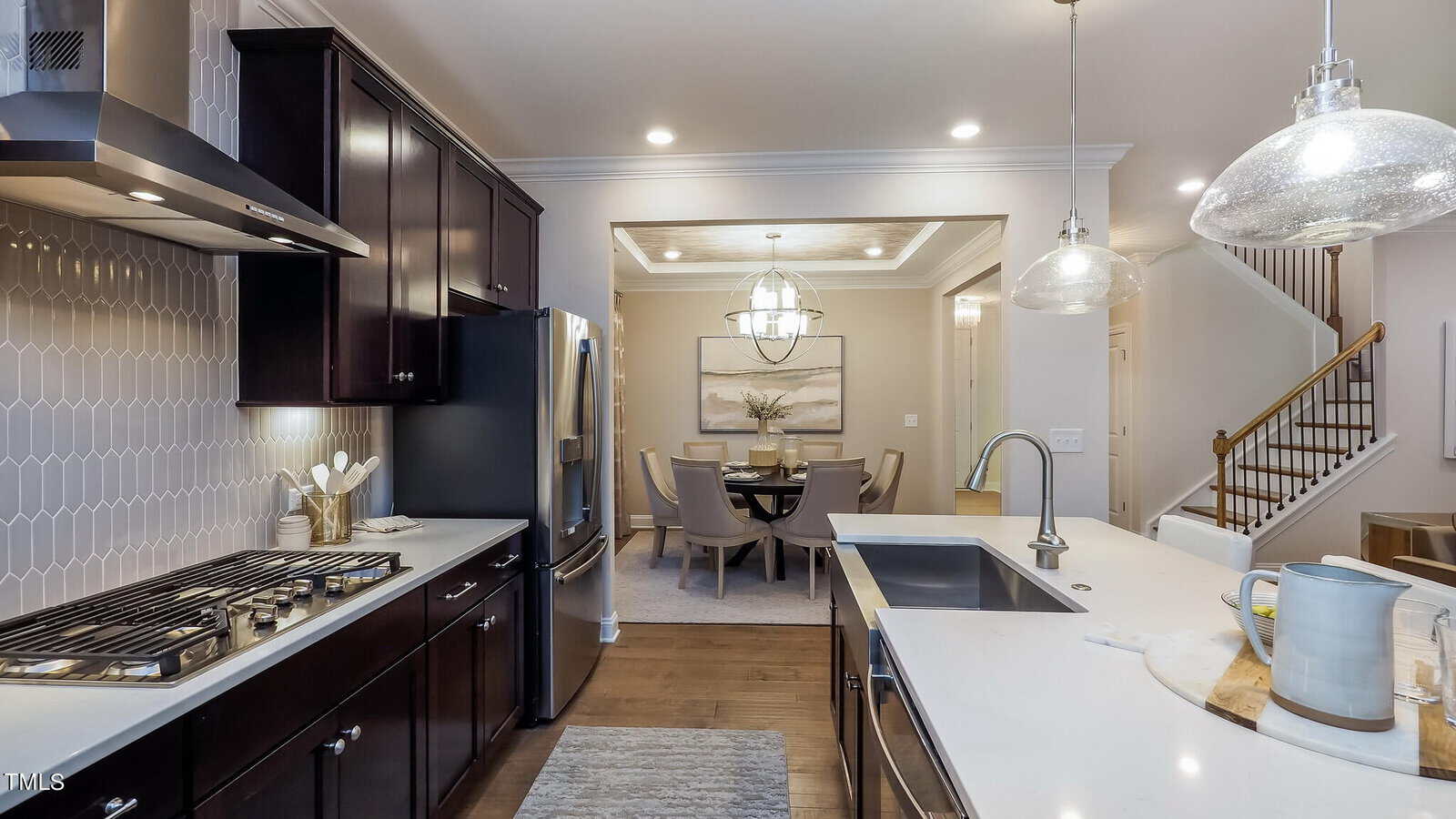 Modern kitchen with dark cabinets, stainless steel appliances, and a white island with a sink. Open layout connects to a dining area with a round table, chairs, and a chandelier. Stairs are visible in the background.