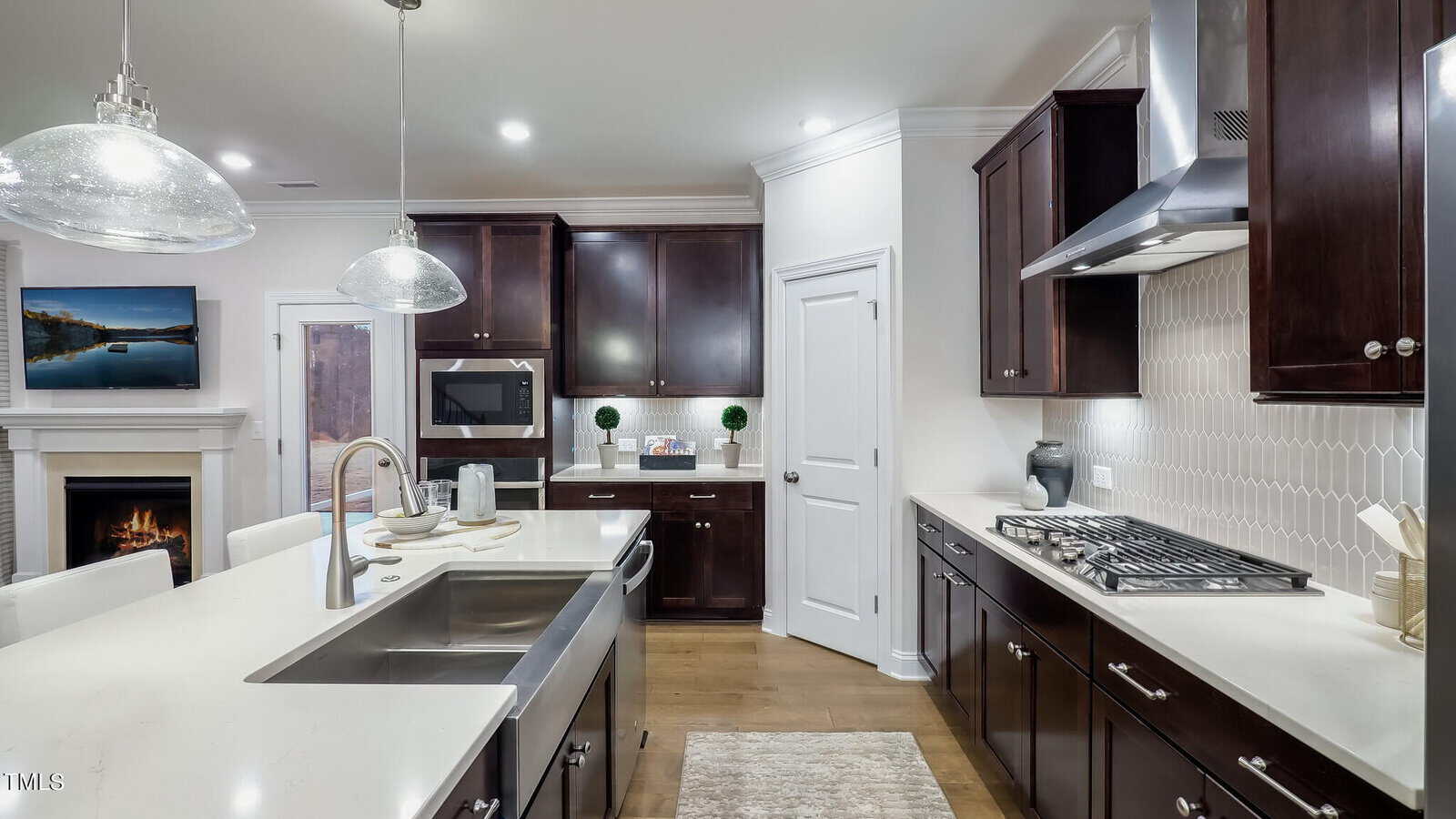 Modern kitchen with dark wood cabinets, white countertops, a large island with a double sink, stainless steel appliances, gas stovetop, pendant lights, and a view into the living area with a fireplace and TV.