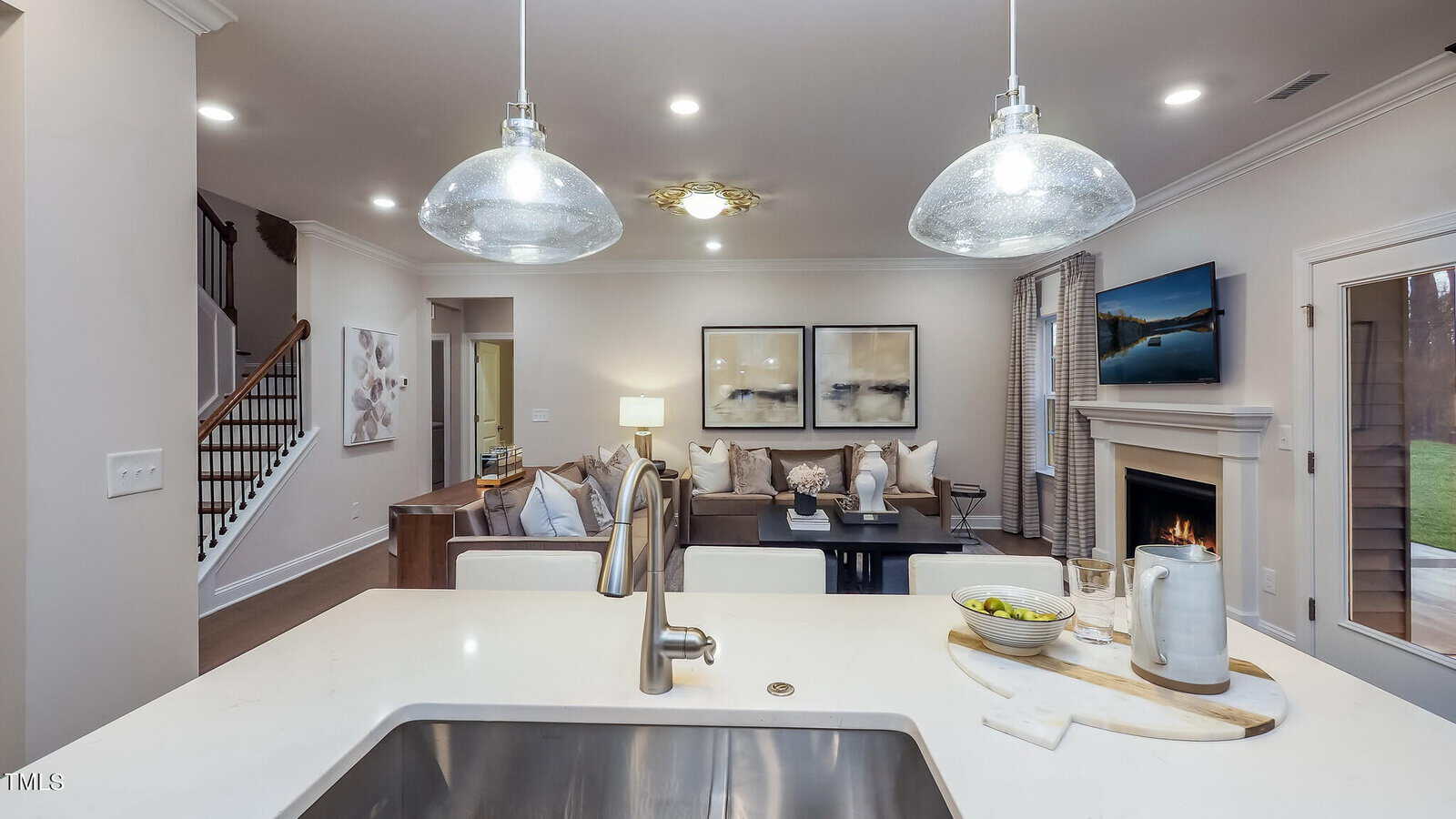 Modern open-concept living space with a kitchen island and sink in the foreground, overlooking a living room with a sectional sofa, wall art, fireplace, and TV. Pendant lights hang above the island.