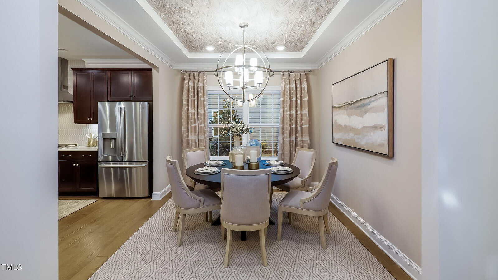 A modern dining room with a round table set for four, light upholstered chairs, a geometric chandelier, large window with curtains, abstract artwork, and a view of the kitchen with stainless steel appliances.