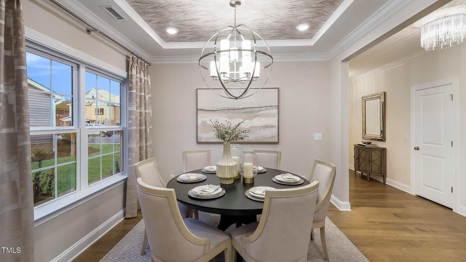 A modern dining room with a round black table set for six, cream-colored chairs, a large vase centerpiece, abstract art on the wall, and a contemporary chandelier overhead. Sunlight streams in through the windows.