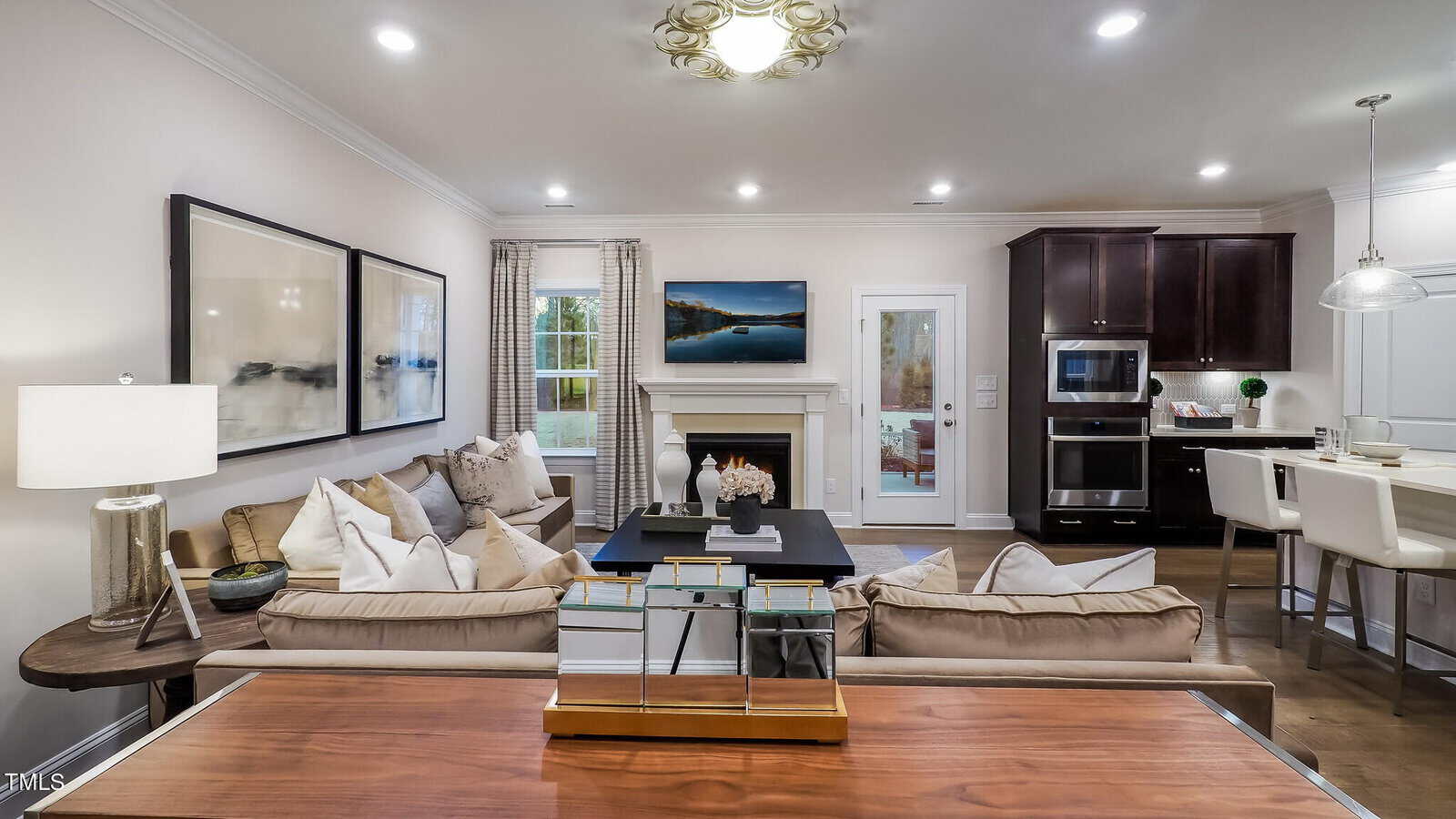 Modern living room with neutral sofas, art on the wall, a fireplace, and a TV. An open kitchen with white bar stools and pendant lights is visible in the background. Cozy, well-lit space with contemporary decor.