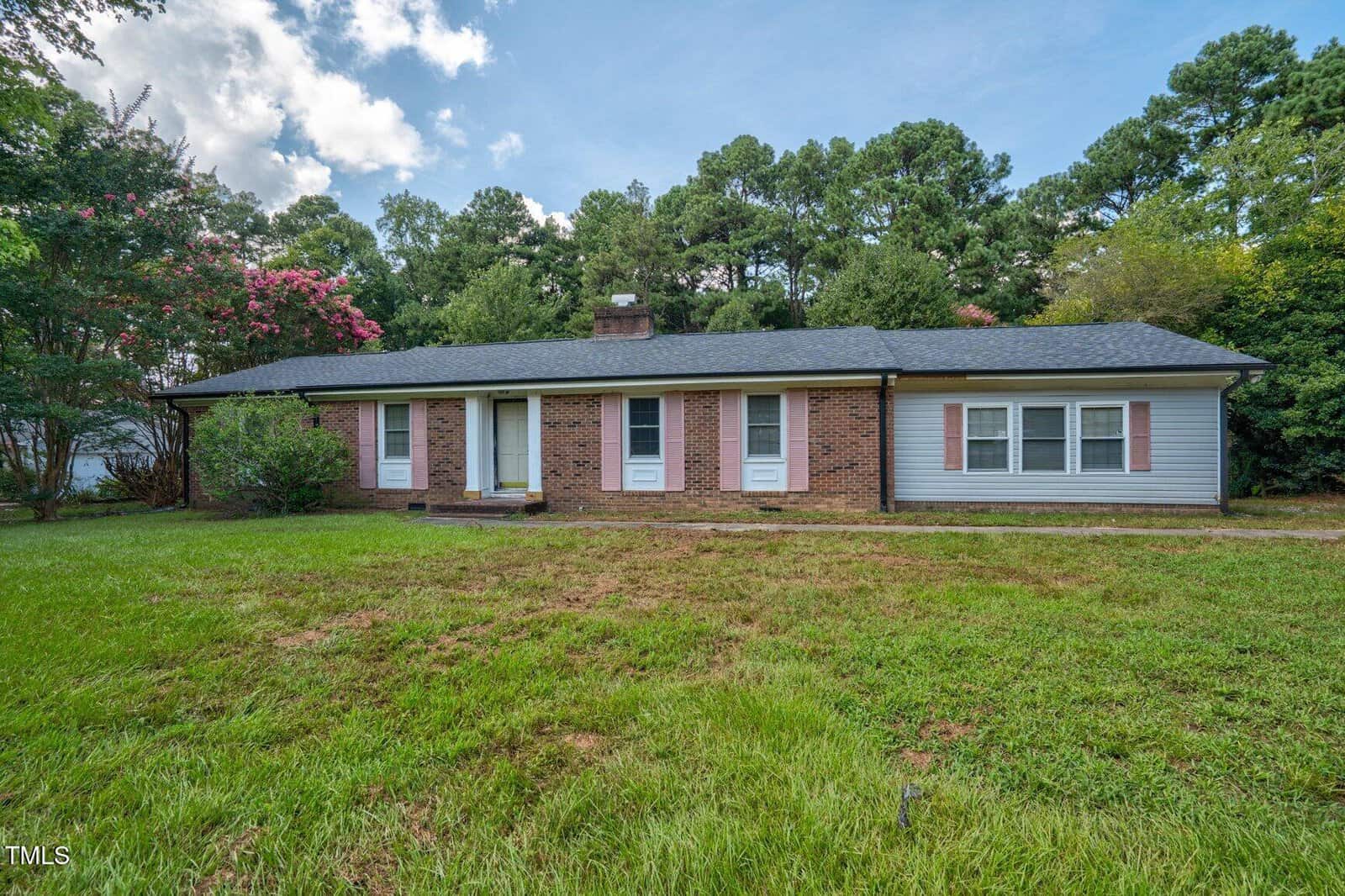 Single-story brick house with white trim, gray shingle roof, and three front doors, set on a grassy yard with trees and bushes in the background under a partly cloudy sky.