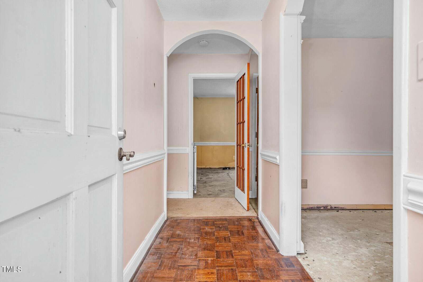 A view into a house hallway with light pink walls, white trim, and wood parquet flooring. The hallway leads to rooms with open doors, revealing bare, partially unfinished or worn floors in adjoining rooms.
