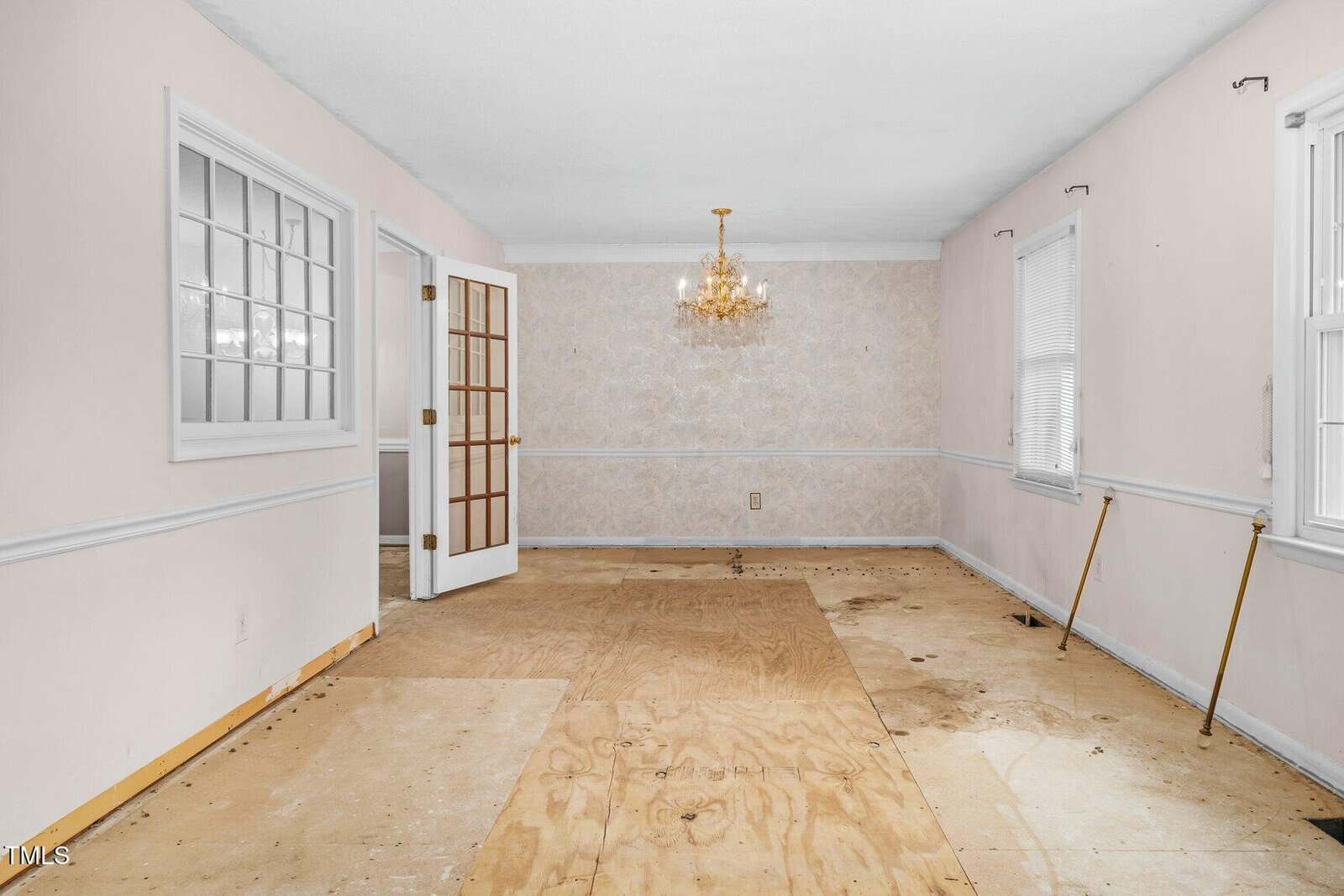 An empty room with light pink walls, white trim, a chandelier, and plywood subfloor. There are two windows, a French door, and some wall damage and stains visible on the floor.