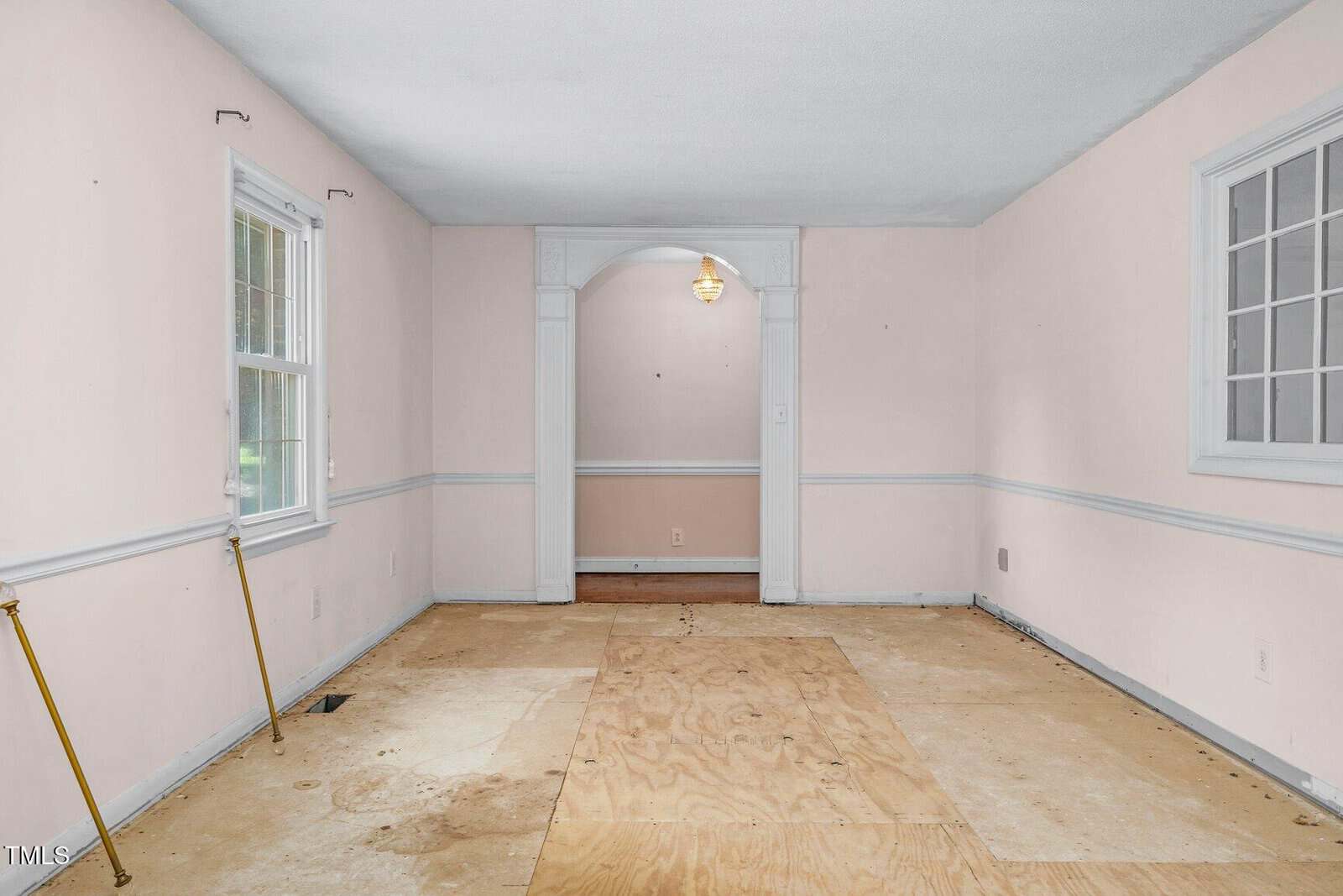 Empty room with light pink walls, white trim, two windows, and exposed plywood floor. Brass curtain rods are attached below the windows, and an archway leads to a small entry area with a hanging light fixture.