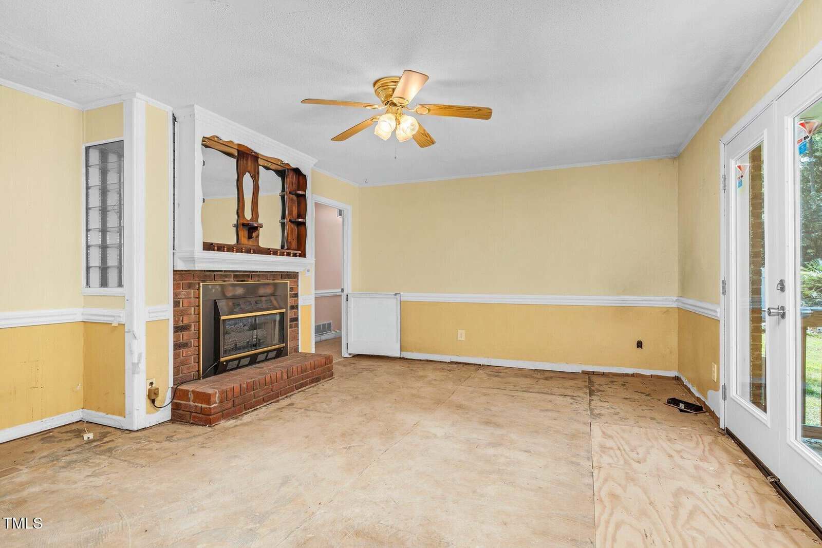 A mostly empty living room with yellow walls, white trim, a brick fireplace with a wooden mantle, a ceiling fan with lights, bare floor, and glass doors leading outside.