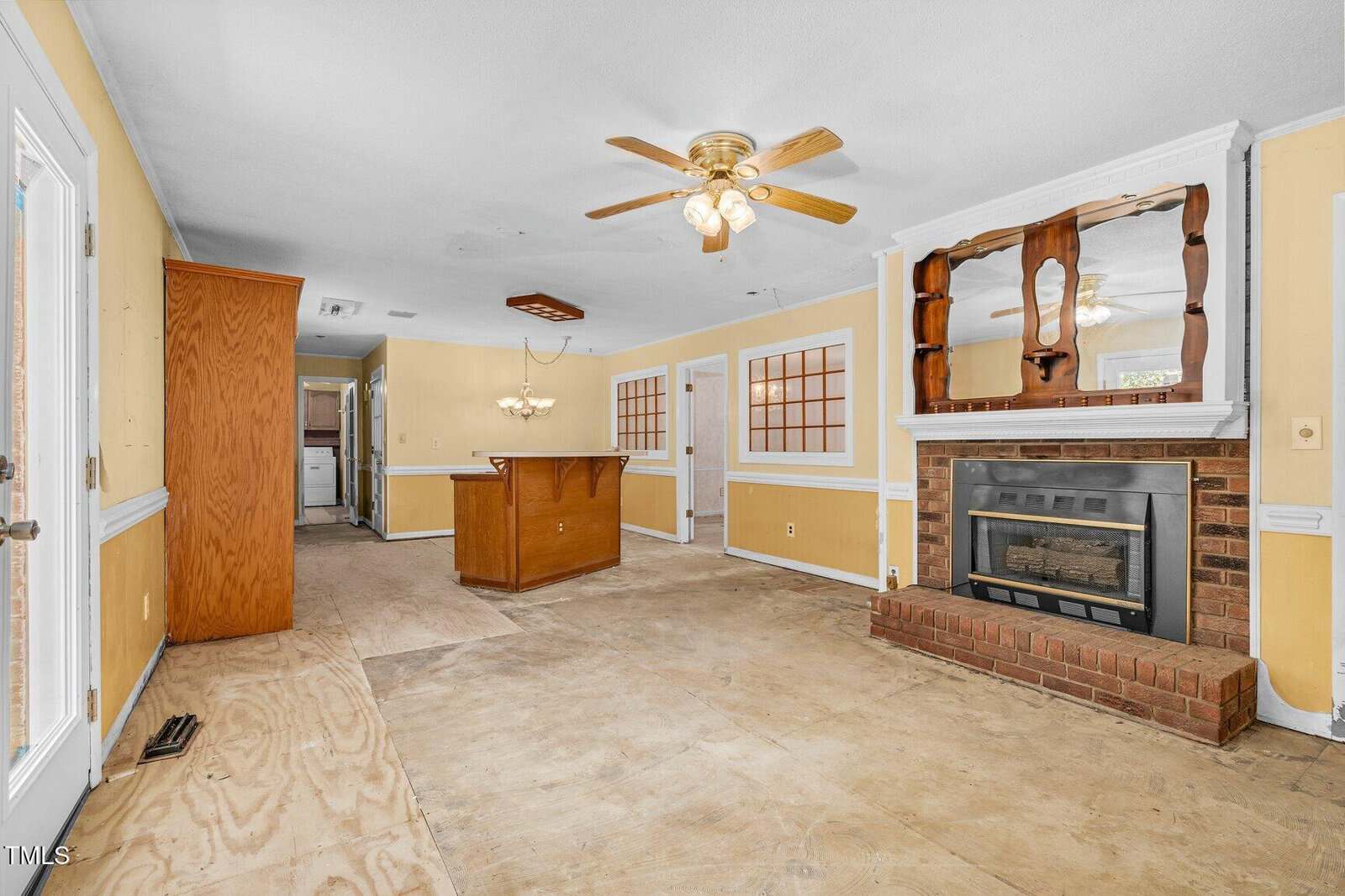Unfinished living area with bare floors, yellow walls, ceiling fan, brick fireplace, wood mantle, and built-in shelving. Open space leads to dining area, kitchen, and another room in the background.