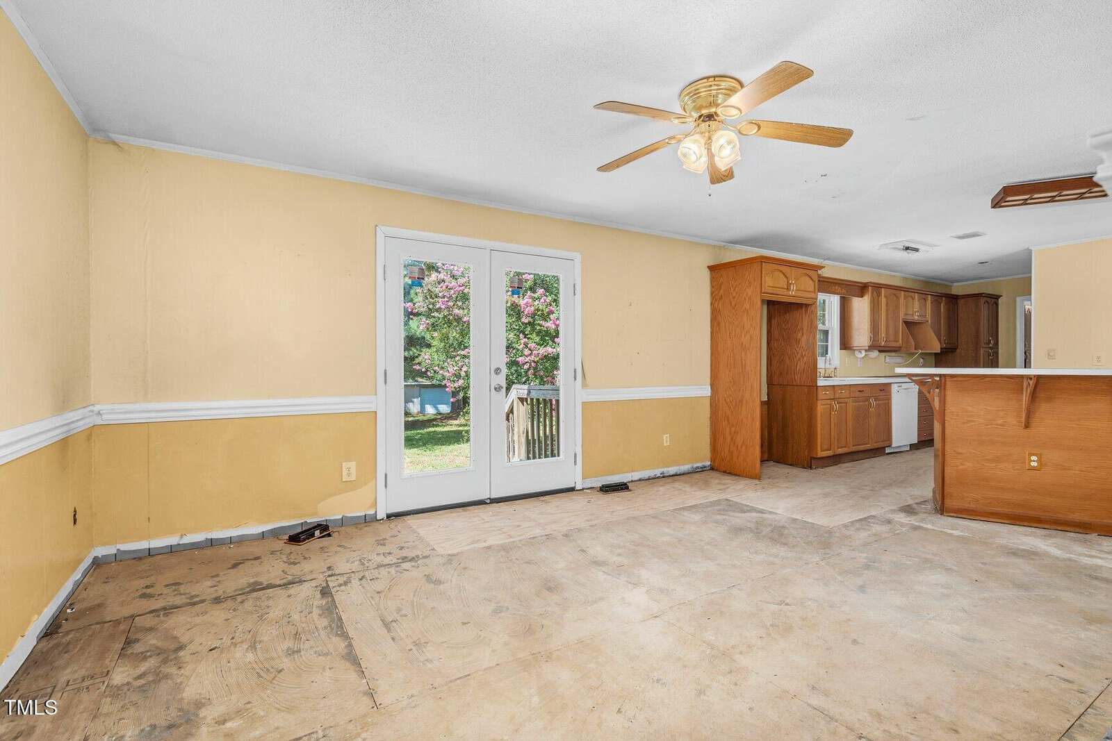 A partially renovated room with bare floors, yellow walls, a ceiling fan with lights, double glass doors leading outside, and an open kitchen with wood cabinets visible in the background.