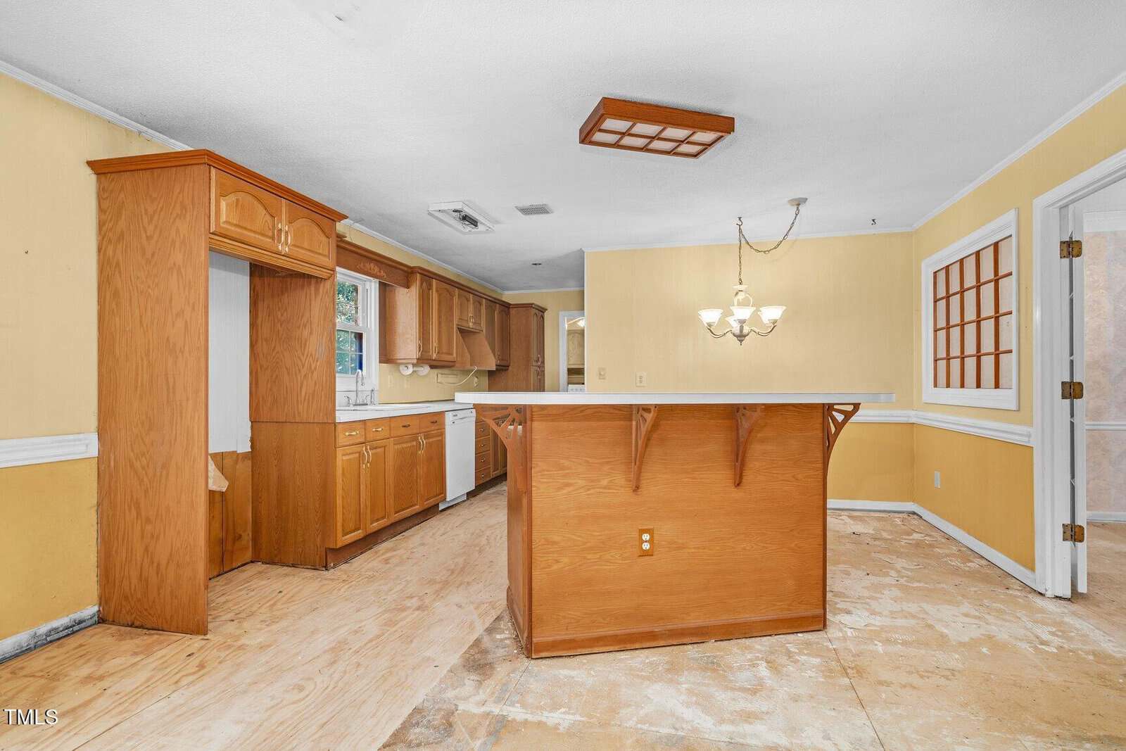 A kitchen with yellow walls, wood cabinets, a large island, white countertops, and no flooring. Appliances are missing; only a dishwasher remains. A chandelier hangs above the island and a window is above the sink.