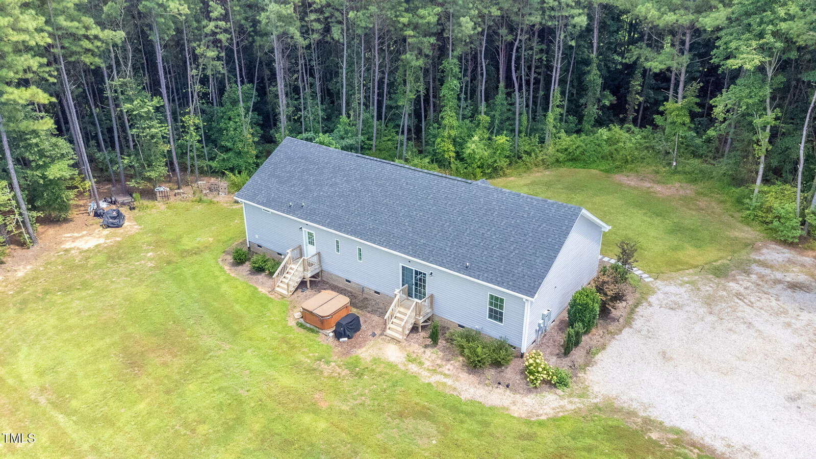 Aerial view of a single-story gray house with a dark roof, two staircases leading to separate doors, a hot tub in the backyard, and surrounded by grass, gravel, and dense trees.