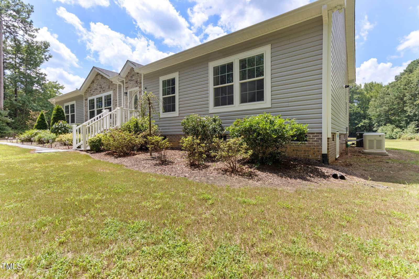 Single-story gray house with white trim and large windows, surrounded by bushes and landscaping. There is a white-railed porch and the house is set on a grassy lawn with trees and blue sky in the background.