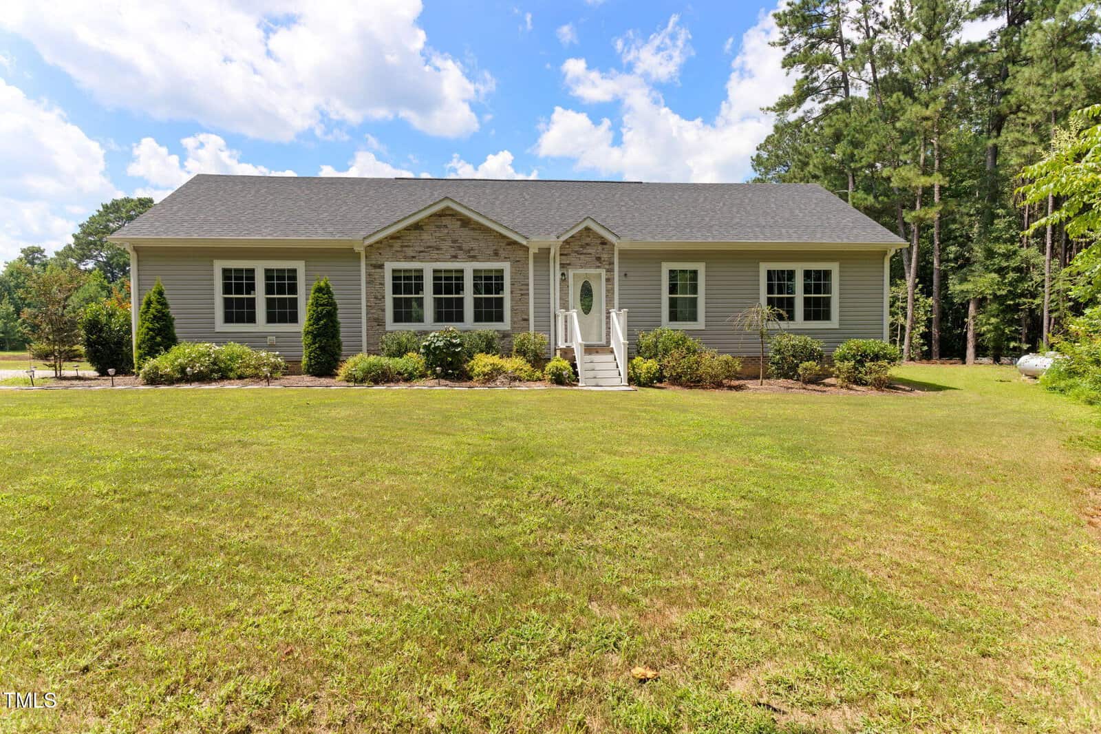 Single-story house with gray siding, a dark shingled roof, and stone accents around the front door. Steps lead to the entrance, and the house is surrounded by lawn, bushes, and trees under a partly cloudy sky.