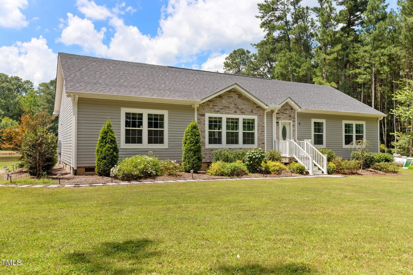 A single-story house with light gray siding, stone accents, white trim, and a small front porch with white railings, surrounded by landscaped bushes and trees, set against a green lawn and forested background.