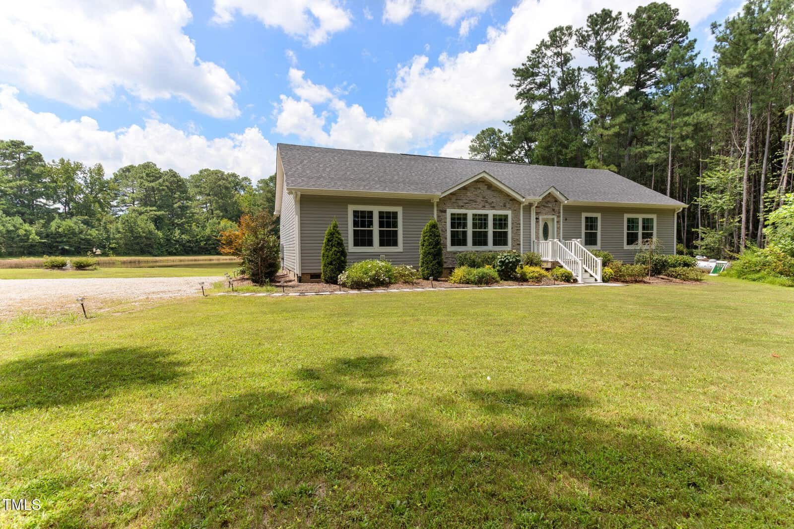 A single-story light gray house with white trim, surrounded by a large green lawn, trees in the background, and a partly cloudy sky above. Steps lead to the front porch with shrubs lining the walkway.