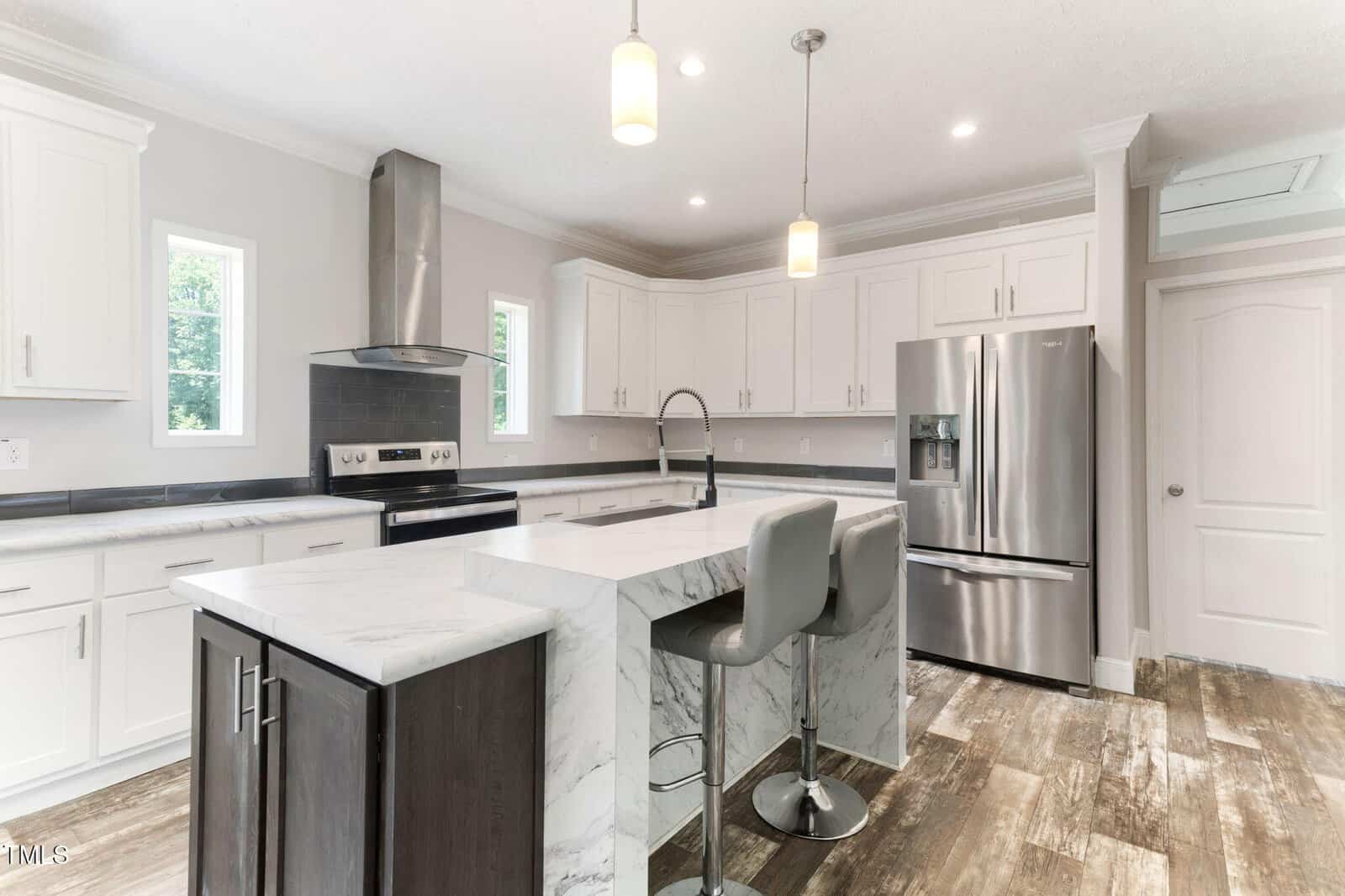 Modern kitchen with white cabinets, marble island with seating, stainless steel appliances, pendant lights, and wood-look tile flooring. Two small windows provide natural light.