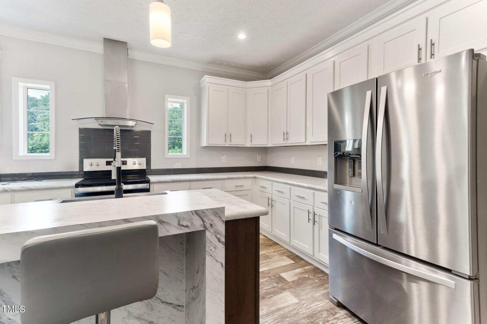 Modern kitchen with white cabinets, stainless steel appliances, a marble island with a gray barstool, electric stove, large refrigerator, and two small windows above the countertops letting in natural light.