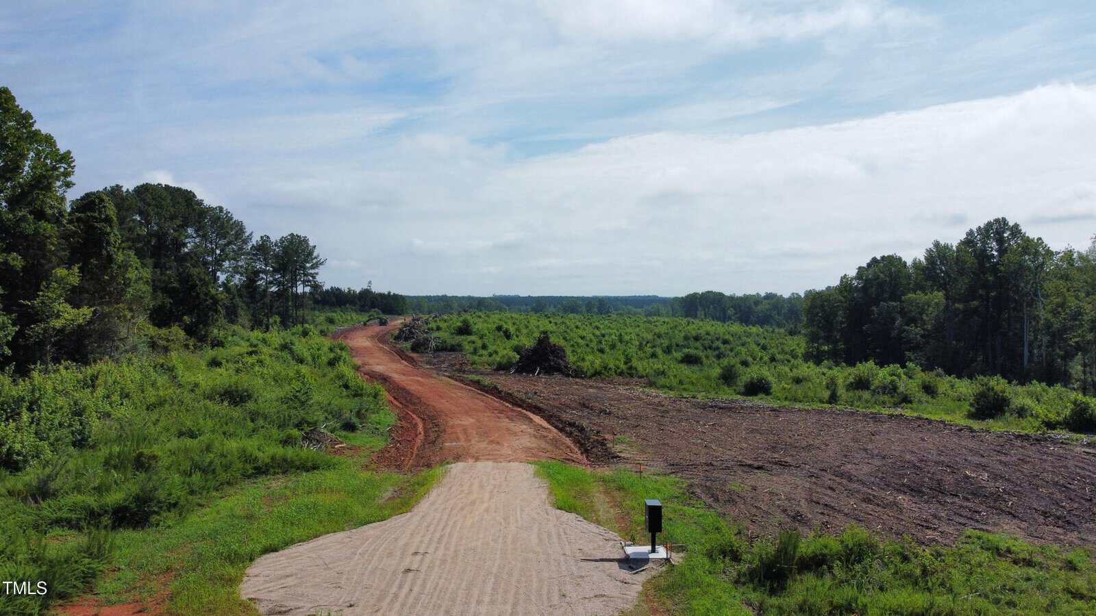 A dirt road leads through a cleared area with some piles of debris, surrounded by green trees and bushes under a partly cloudy sky.