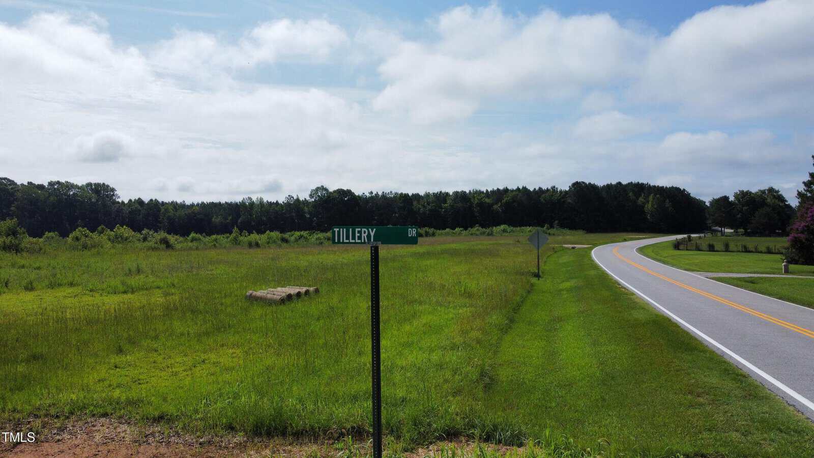 A rural road curves past a grassy field and a street sign labeled "TILLERY DR." Trees line the horizon under a partly cloudy sky.