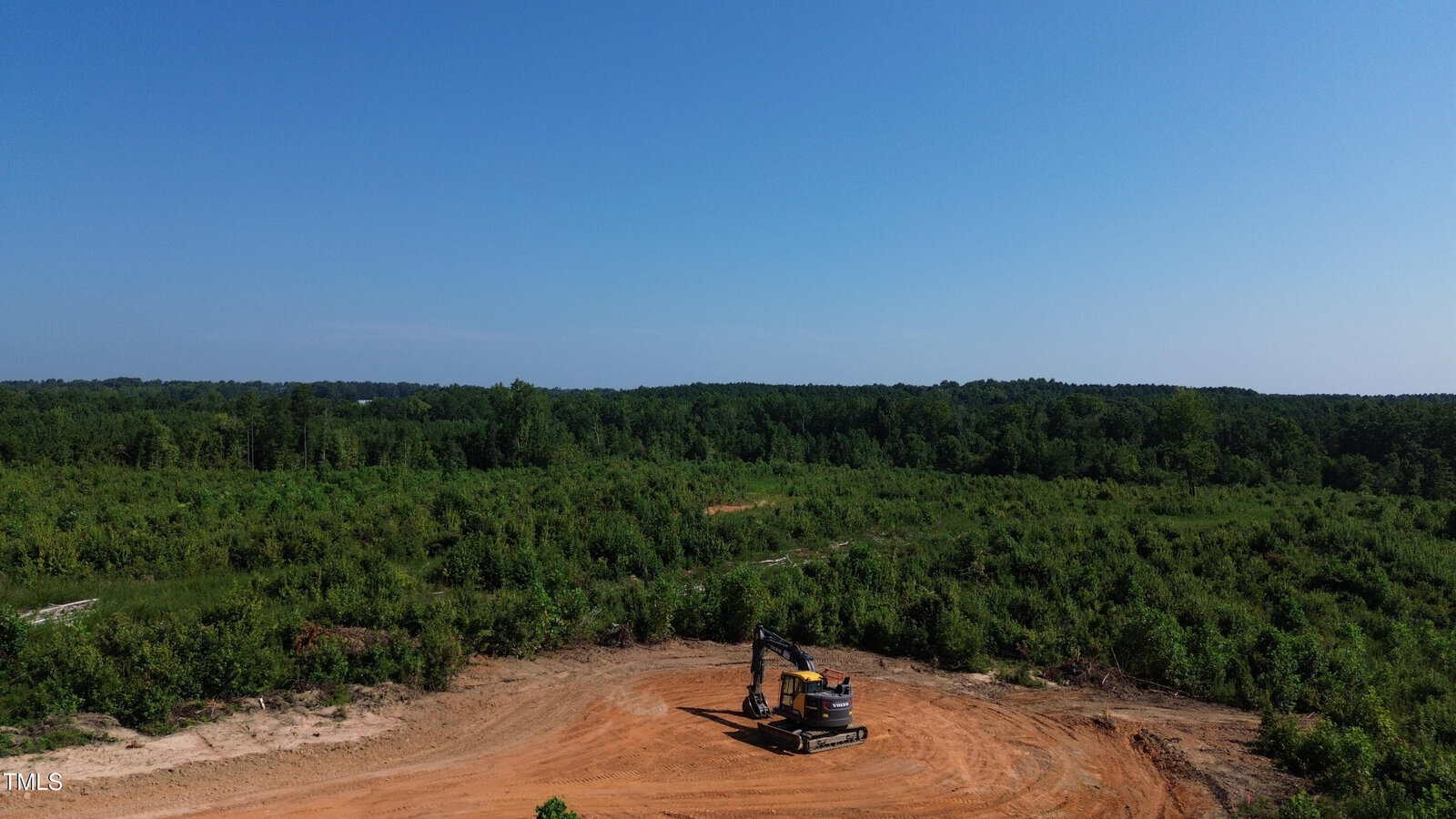 An excavator sits on a cleared dirt area surrounded by green vegetation and dense forest under a clear blue sky.