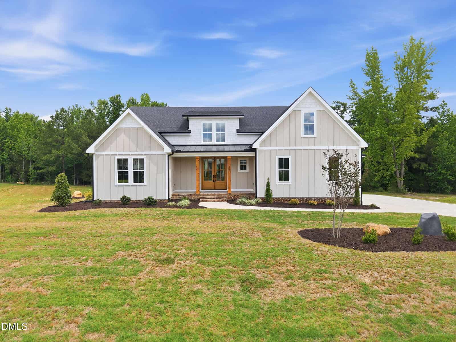 A modern farmhouse-style home with light-colored vertical siding, a dark roof, and wood-framed double front doors. The house is surrounded by a large, neatly manicured lawn and bordered by trees under a blue sky.
