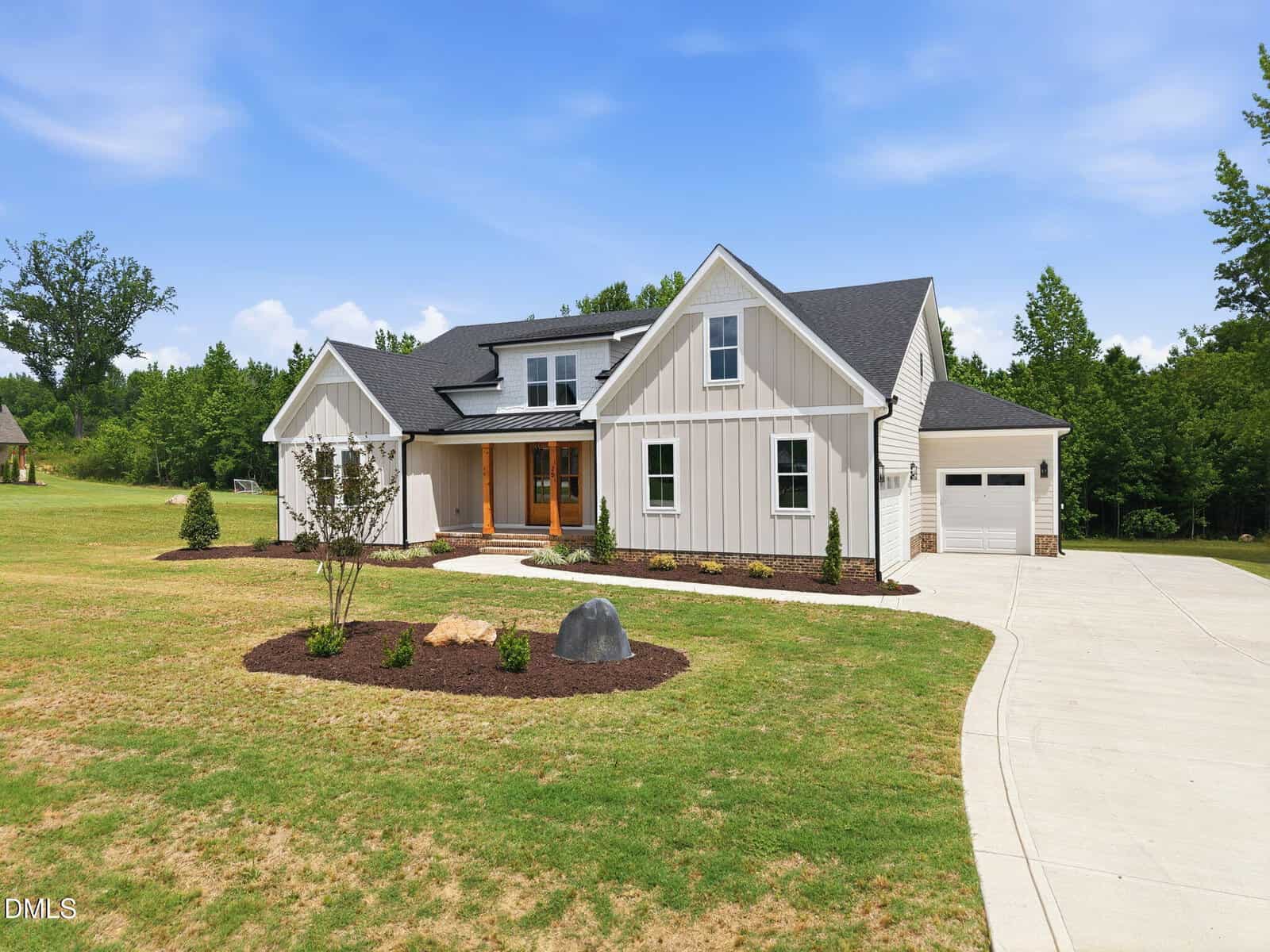 A modern, white farmhouse-style home with dark roof shingles, large front porch, manicured lawn, and landscaped garden beds. A curved driveway leads to an attached garage. Trees and blue sky are in the background.