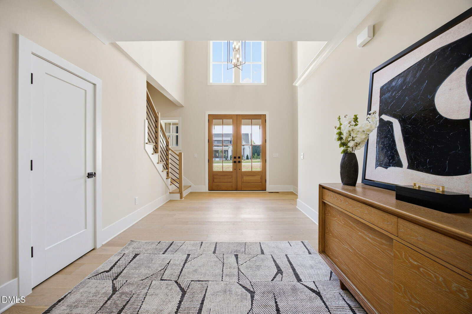 Bright, modern entryway with tall ceilings, light wood floors, double wooden front doors, a staircase to the left, a large abstract painting above a wooden console, and decorative flowers on the console.