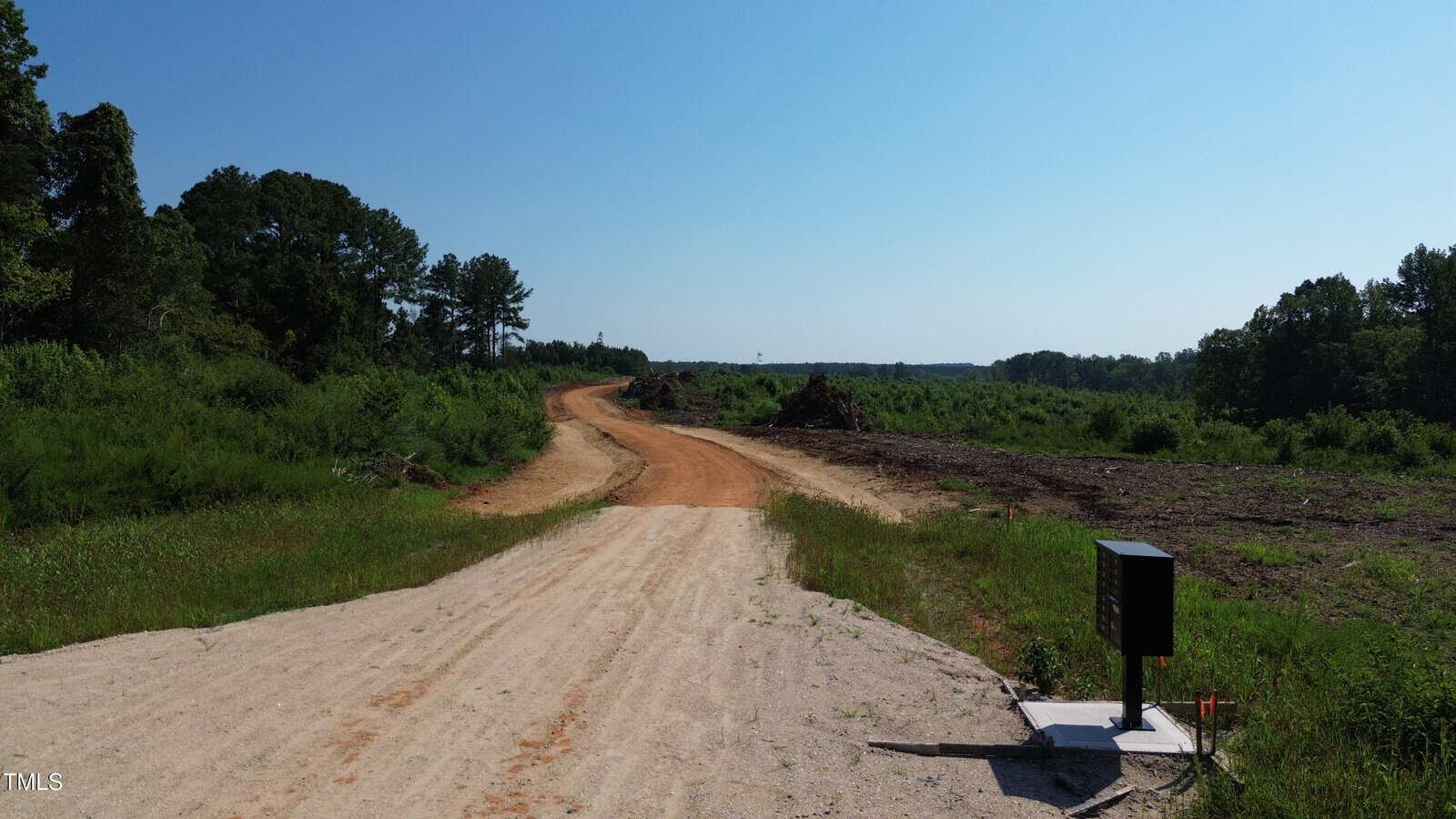 A dirt road stretches through a cleared, grassy area with trees in the distance under a clear blue sky. A black mailbox stands on the right side near the road's edge.