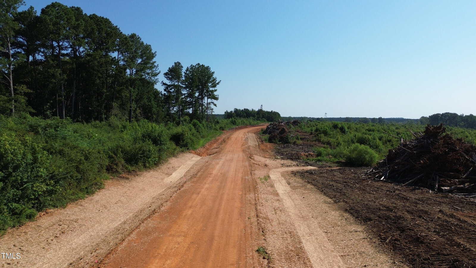 A dirt road runs through a cleared, rural area bordered by green trees and bushes on the left and piles of brush and debris on the right, beneath a clear blue sky.