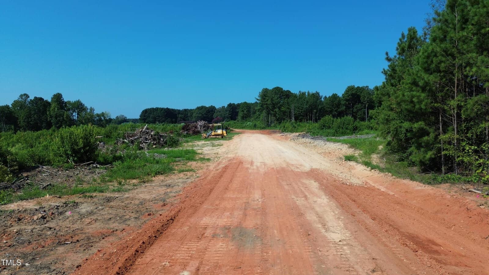 A dirt road stretches through a cleared area with trees and bushes on each side. Construction equipment is visible in the distance under a clear blue sky.