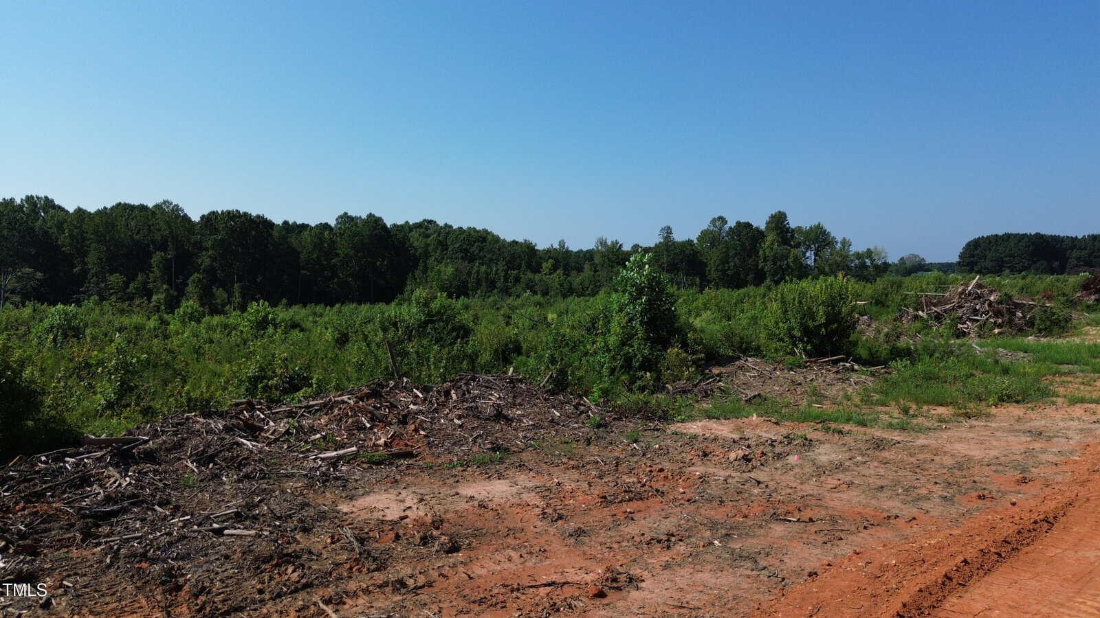 A cleared plot of land with piles of branches and tree debris in the foreground, surrounded by green trees and bushes under a clear blue sky.