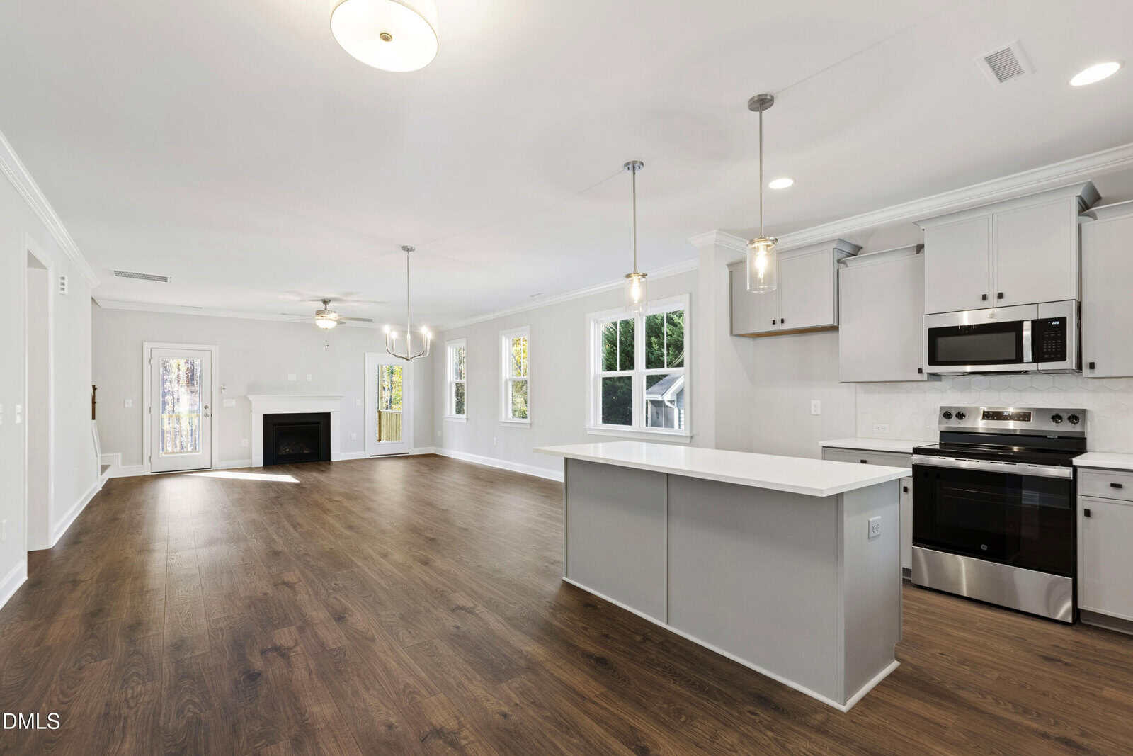 Open-concept kitchen and living room with wood floors, white walls, large windows, modern light fixtures, a center island, stainless steel appliances, and a fireplace in the corner.