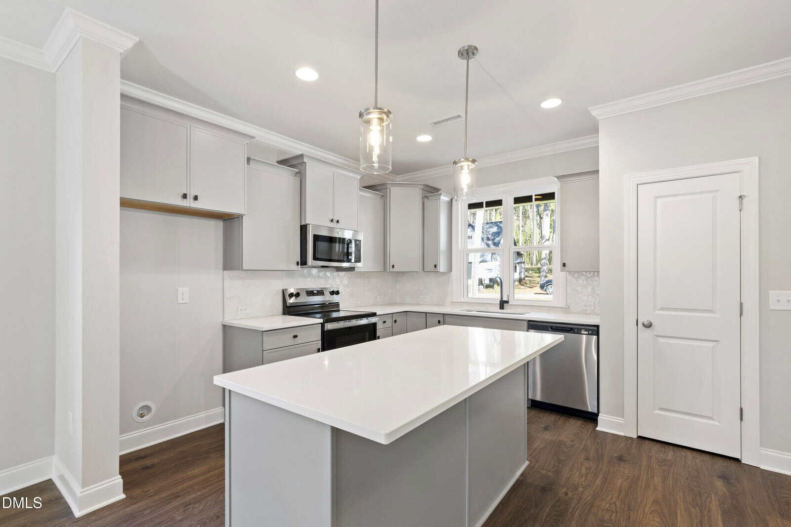 Modern kitchen with white cabinets, stainless steel appliances, a large white island, pendant lights, and a window above the sink. The room features wood flooring and neutral-colored walls.