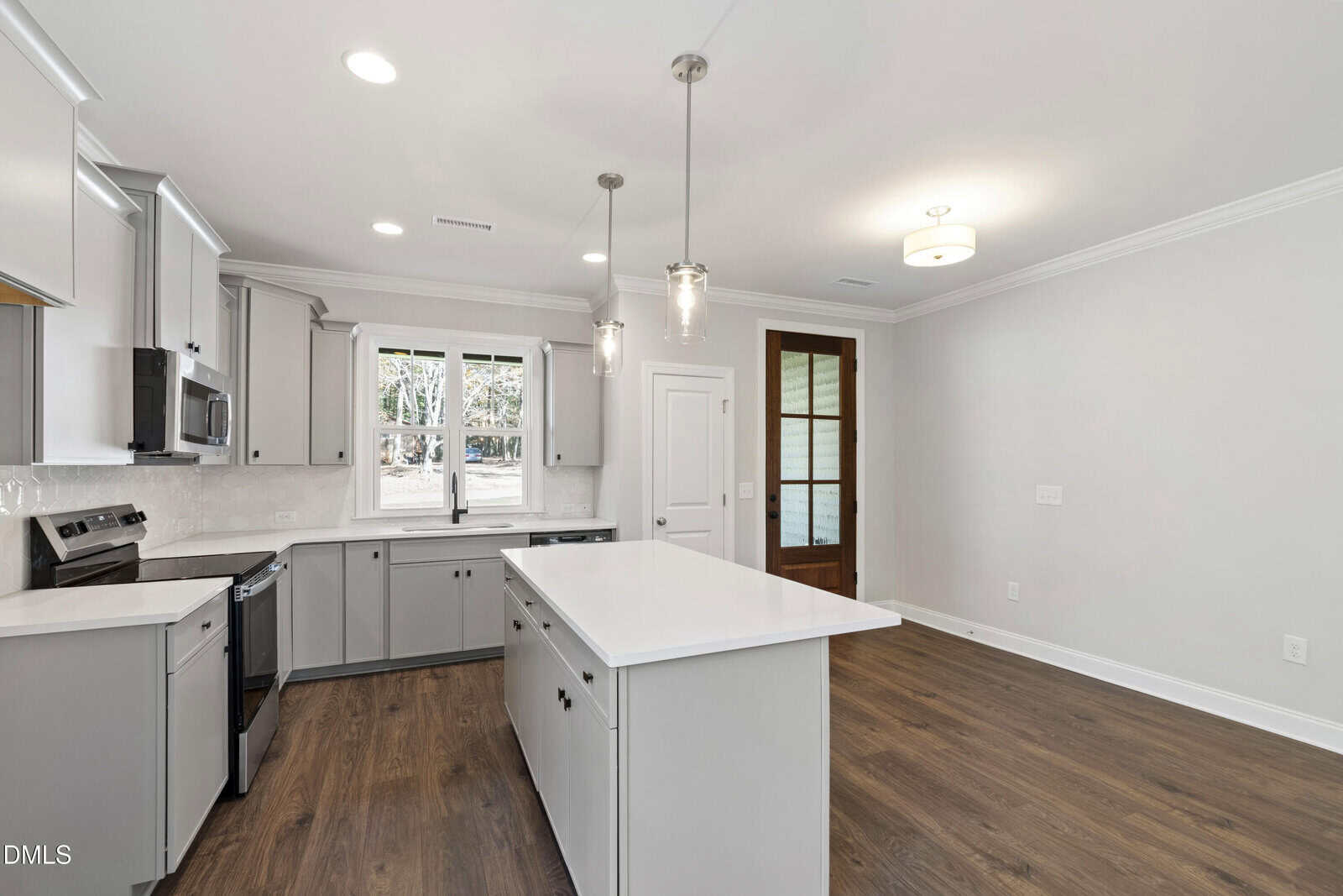 A modern kitchen with gray cabinets, stainless steel appliances, a white island countertop, pendant lights, wood flooring, and a large window over the sink. An adjoining room features double glass doors and white walls.