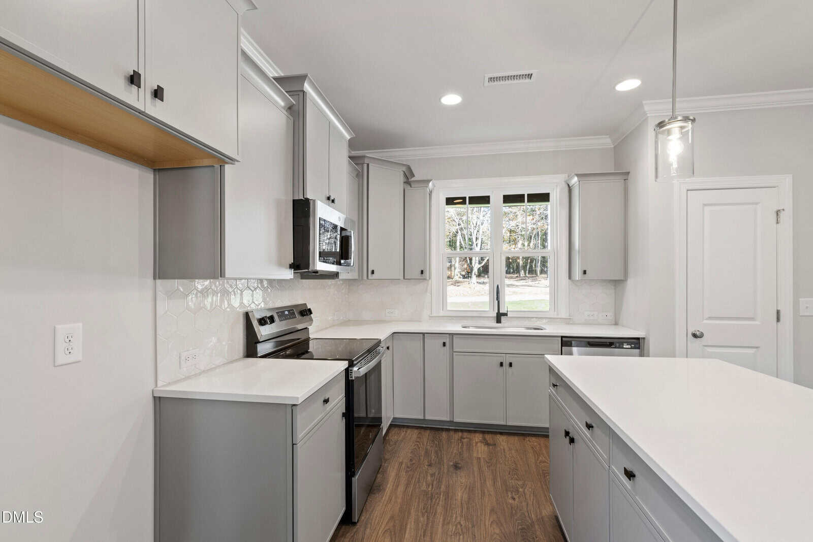 Modern kitchen with light gray cabinets, stainless steel appliances, white countertops, a center island, wood flooring, and a window above the sink letting in natural light. White hexagonal backsplash tiles line the wall.