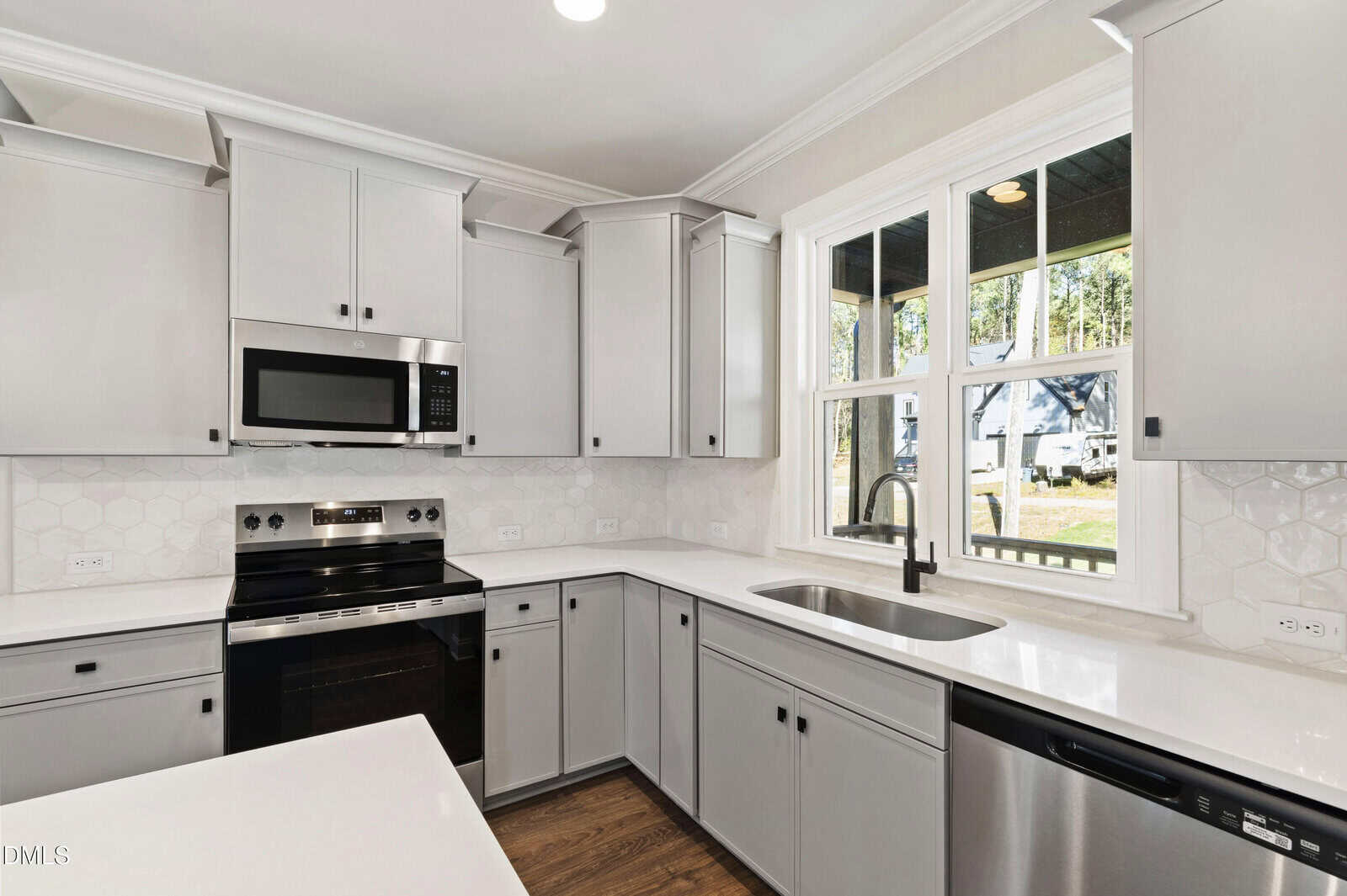 Modern kitchen with light gray cabinets, stainless steel appliances, a black faucet, white hexagonal tile backsplash, and large window above the sink letting in natural light.