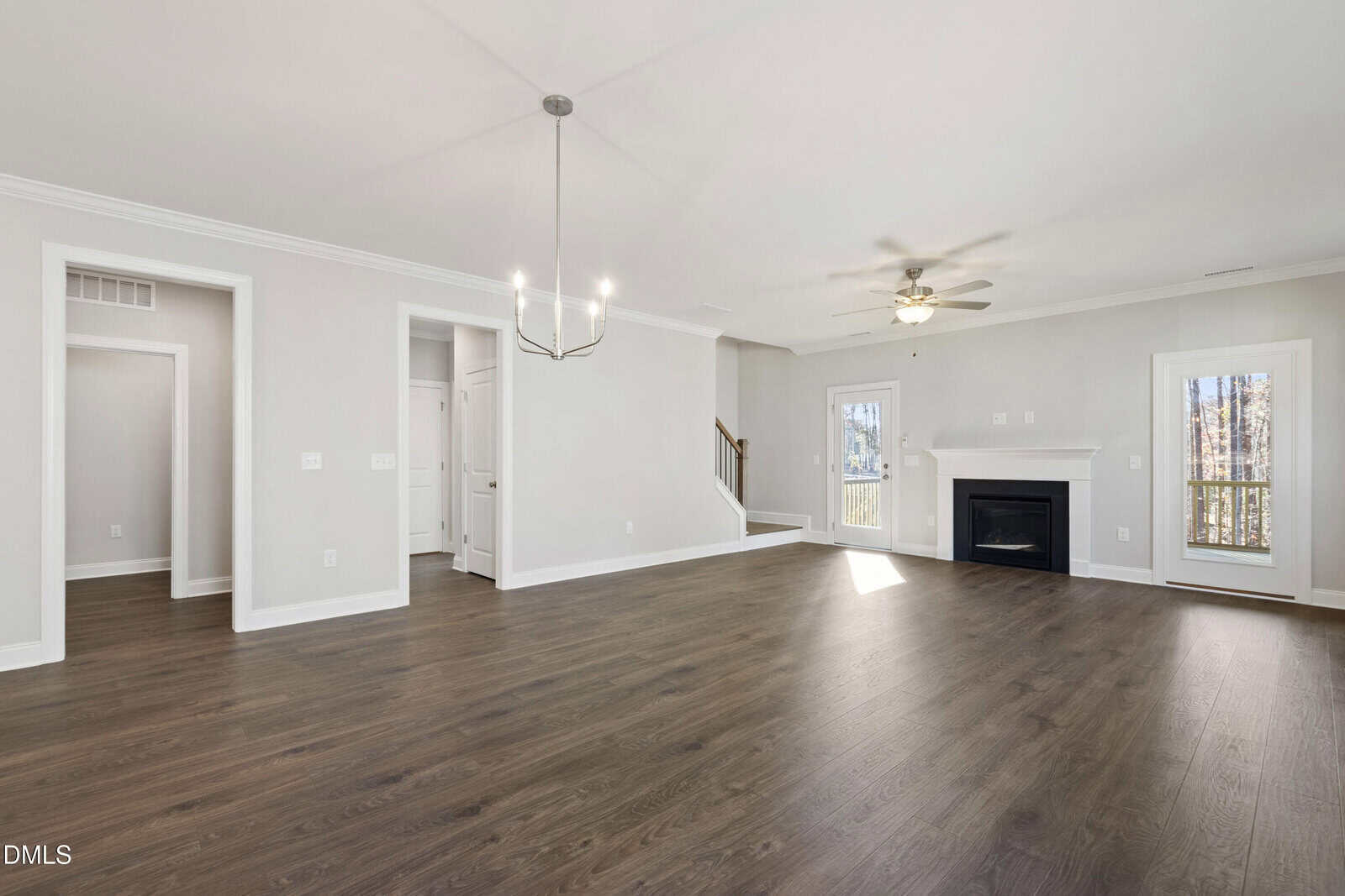 Spacious, empty living room with dark wood floors, light gray walls, modern chandelier, ceiling fan, fireplace, and large windows letting in natural light.