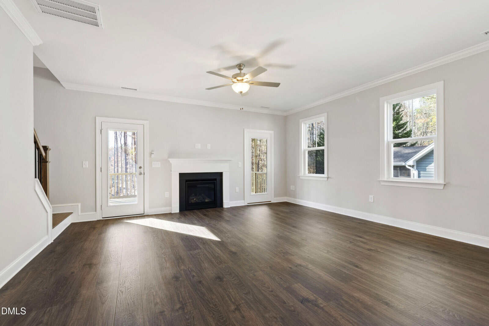 Spacious, empty living room with light gray walls, dark wood flooring, a white ceiling fan, fireplace, several windows, and a glass door leading outside. Stairs are visible on the left side of the room.