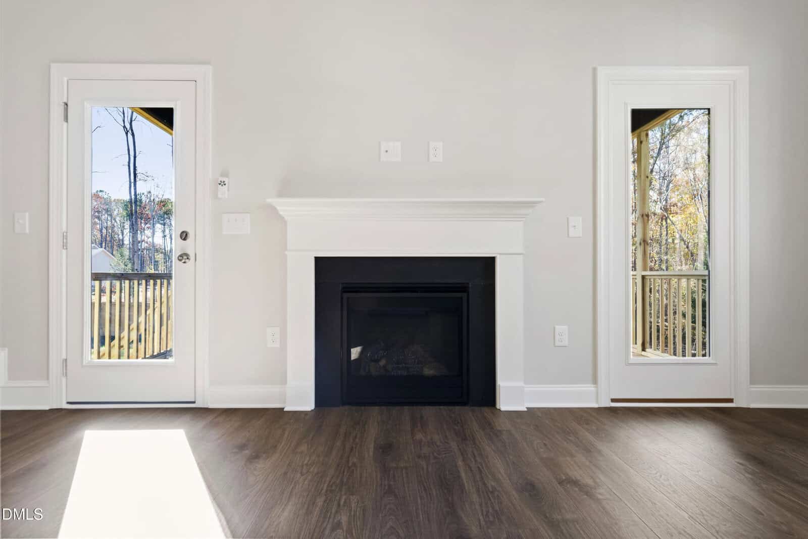 A modern, empty living room with a dark fireplace centered between two tall glass doors, white walls, and wood flooring. Sunlight streams in, and trees are visible outside through the glass doors.