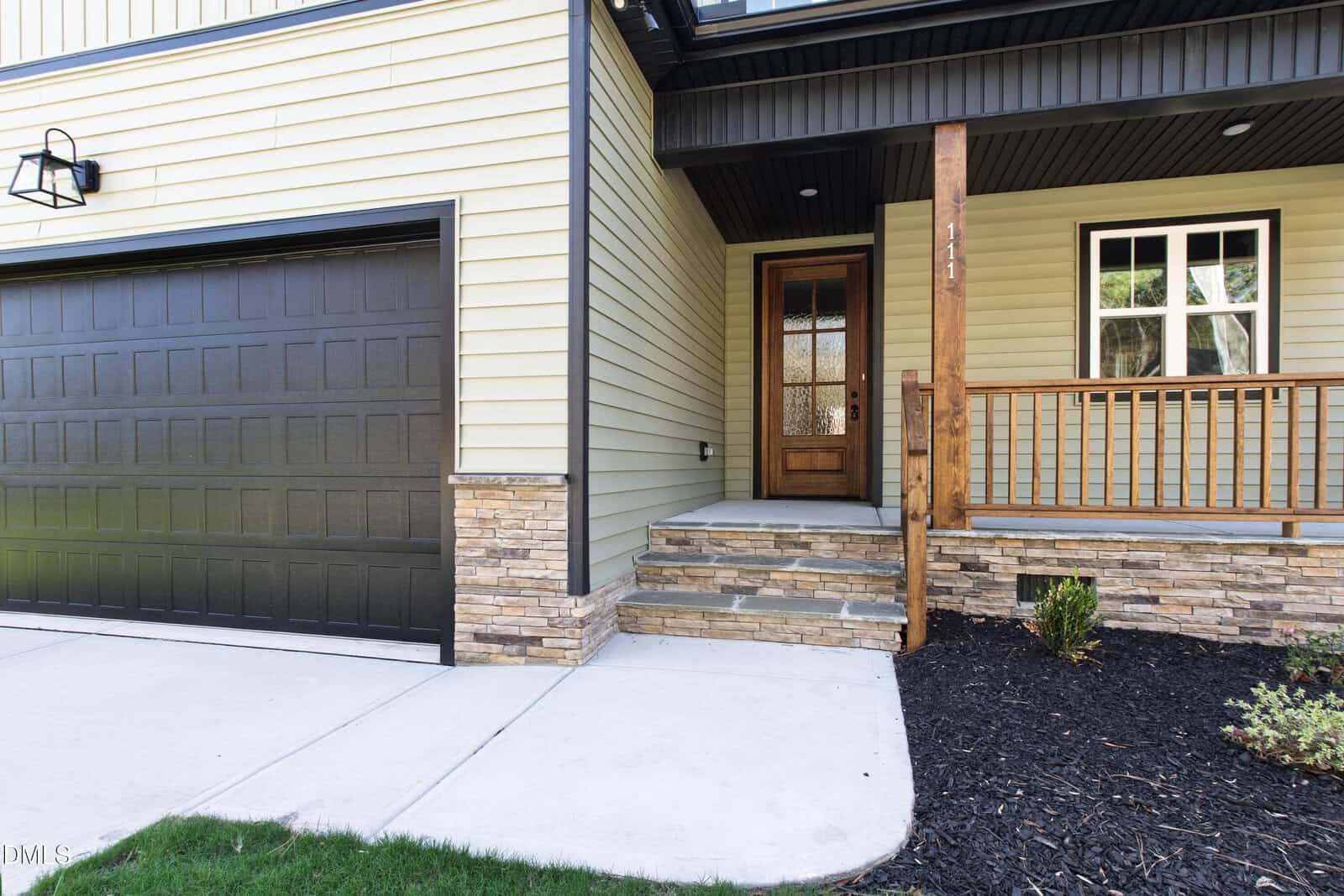 Front view of a house with yellow siding, stone accents, a black garage door, wooden front door, small porch with railing, and concrete steps leading to the entrance. Shrubs and mulch border the walkway.