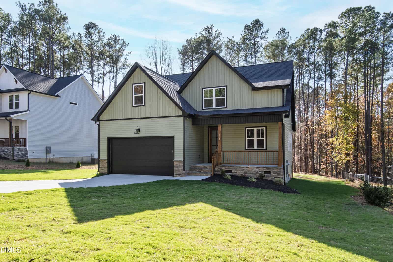 A modern two-story house with green siding, black trim, a double garage, a small front porch, and a well-maintained lawn, set against a backdrop of tall pine trees.