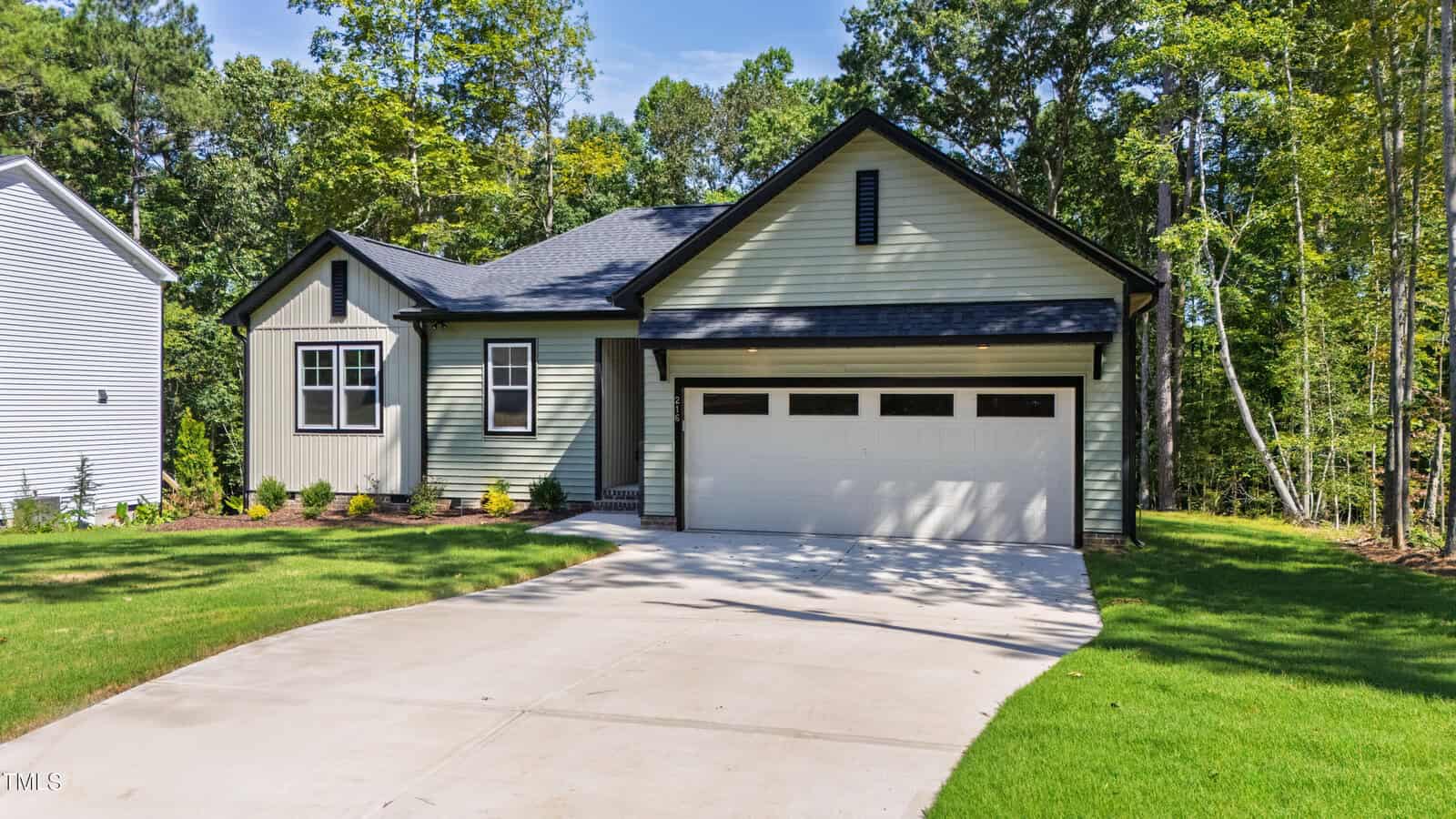 A modern single-story house with light green siding, dark trim, and a two-car garage. The home features a clean driveway, neatly landscaped yard, and is surrounded by tall green trees.