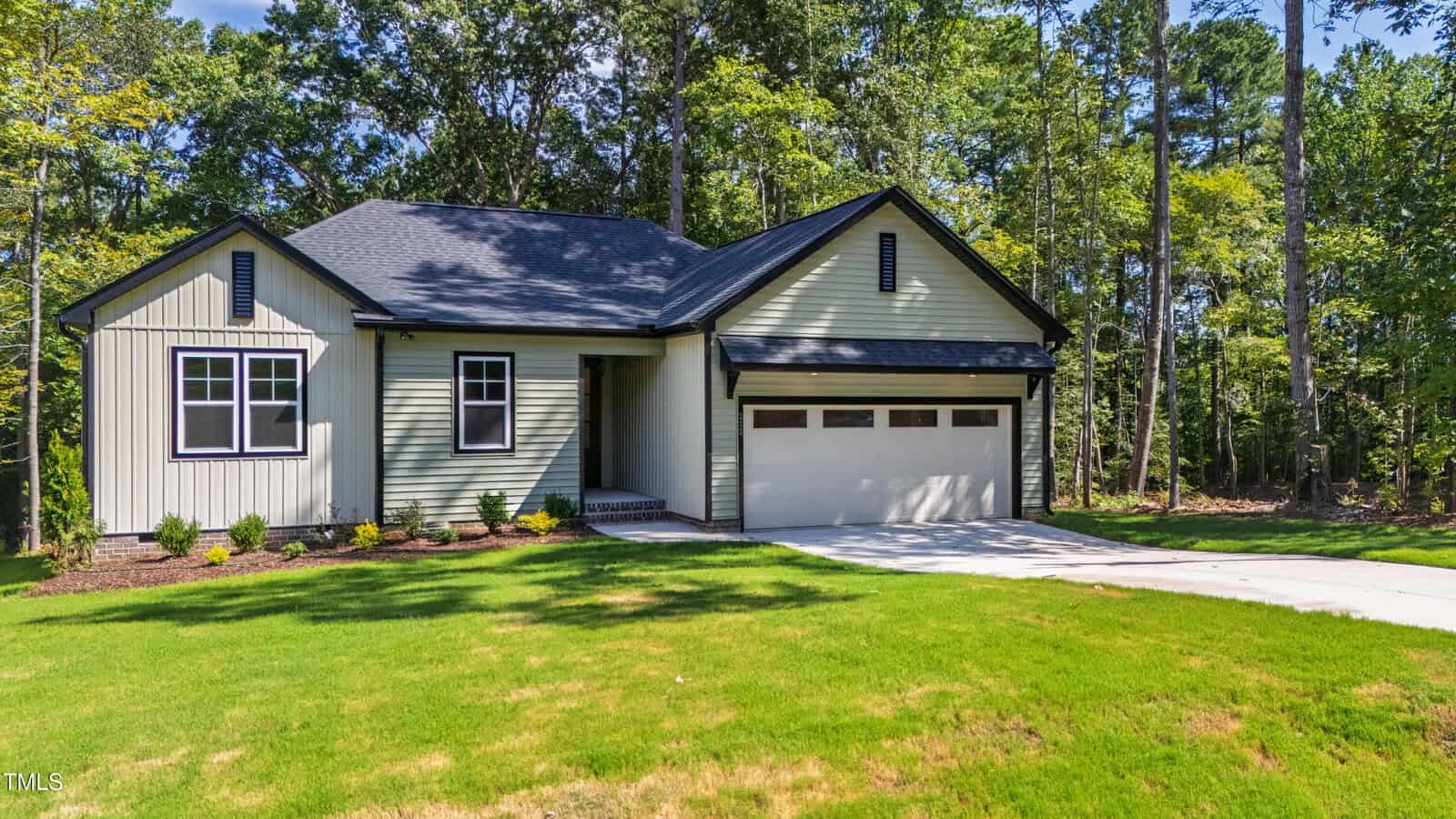 Single-story modern house with light siding, black trim, attached two-car garage, and a covered front entry. Surrounded by a well-kept lawn and trees, the home sits under a clear blue sky.