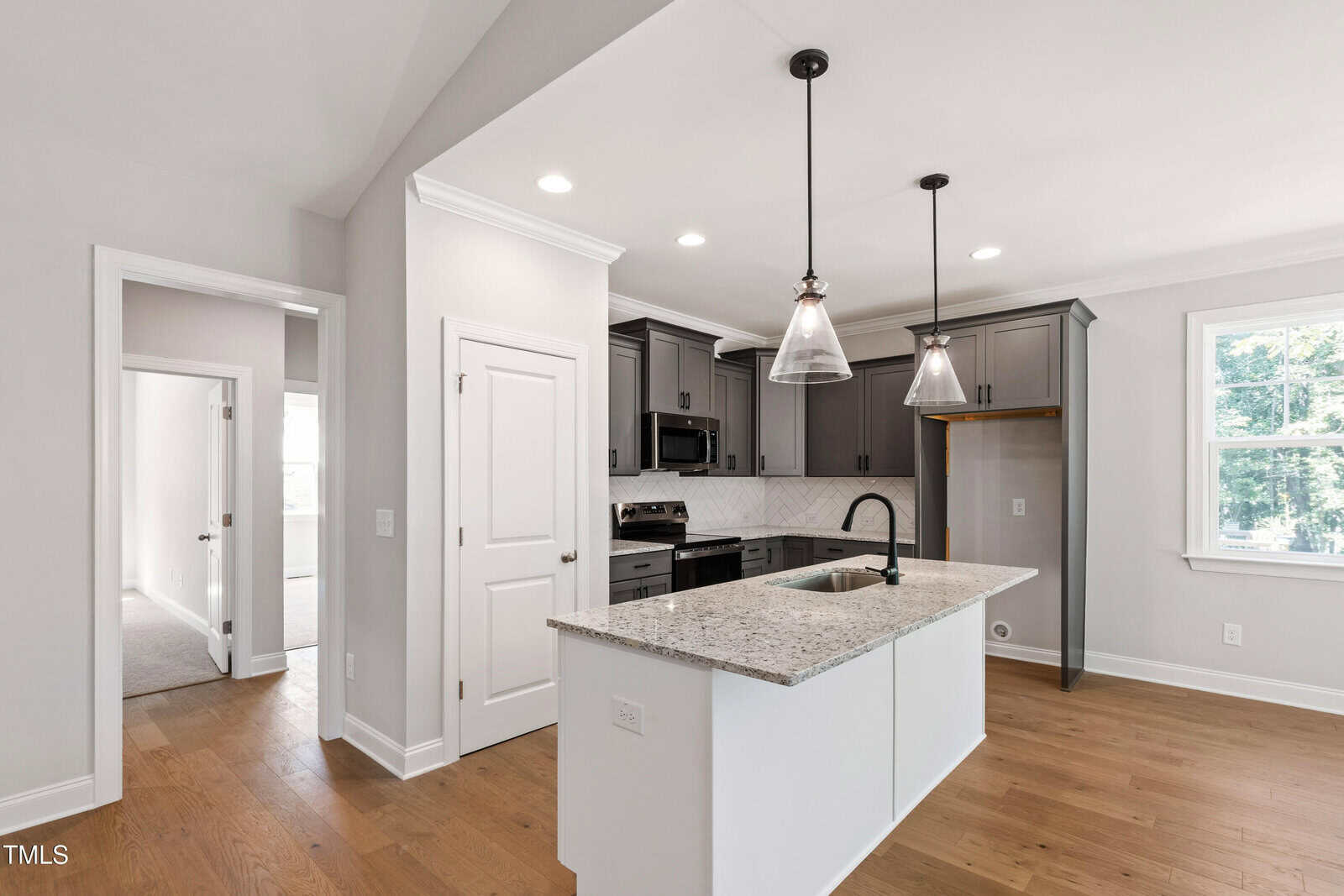 Modern kitchen with gray cabinets, stainless steel appliances, white island with granite countertop, black faucet, two pendant lights, light wood flooring, and white walls. Window lets in natural light. Doorways lead to other rooms.