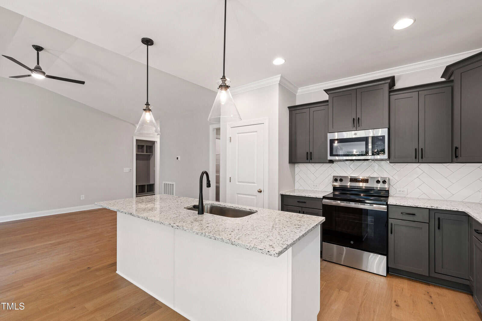 Modern kitchen with a white island, black fixtures, granite countertops, gray cabinets, stainless steel appliances, white tile backsplash, and pendant lighting; open layout to a living area with light wood floors and a ceiling fan.