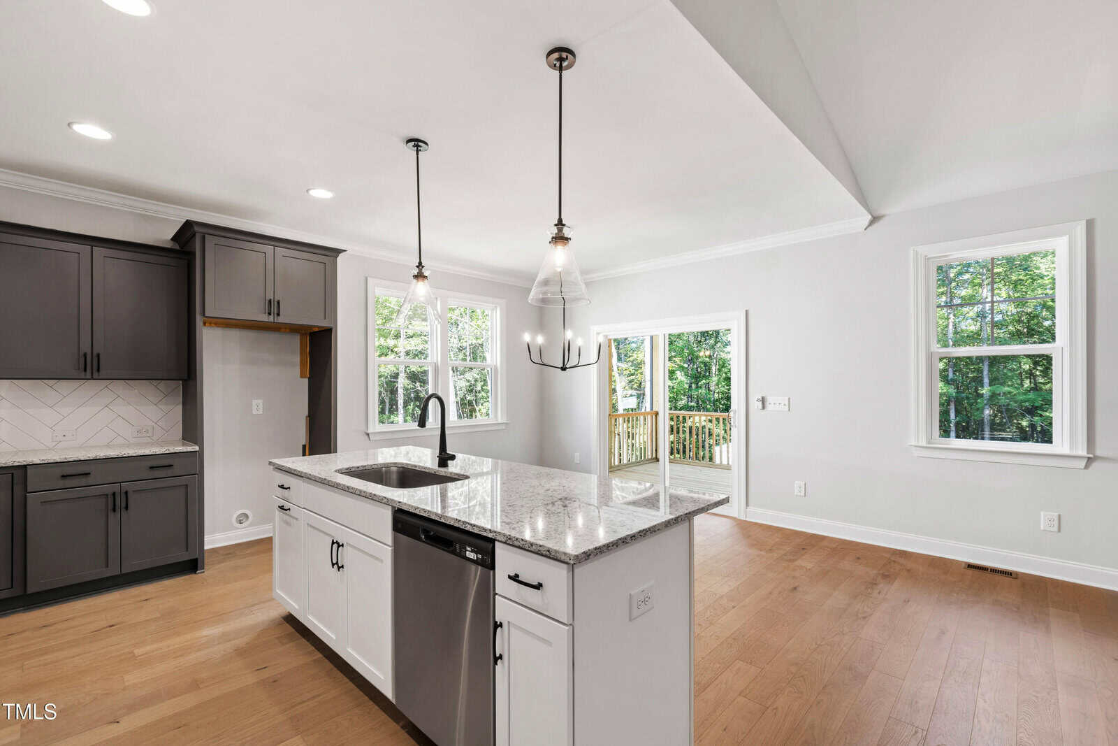 Modern kitchen with a central island featuring a sink and dishwasher, dark and white cabinets, light wood flooring, pendant lighting, large windows, and a sliding glass door leading to a deck with a wooded view.