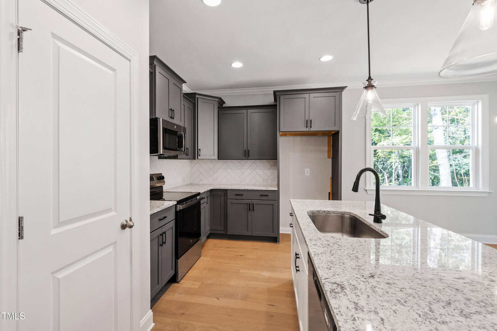 Modern kitchen with gray cabinets, stainless steel appliances, white herringbone backsplash, granite countertops, an island with a sink and black faucet, light wood floors, and large windows letting in natural light.