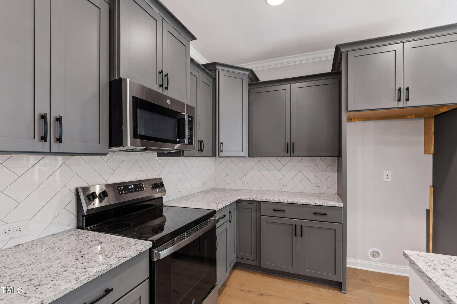 Modern kitchen with gray cabinets, granite countertops, stainless steel oven and microwave, white herringbone tile backsplash, and light wood flooring. An empty cabinet space is visible on the right.